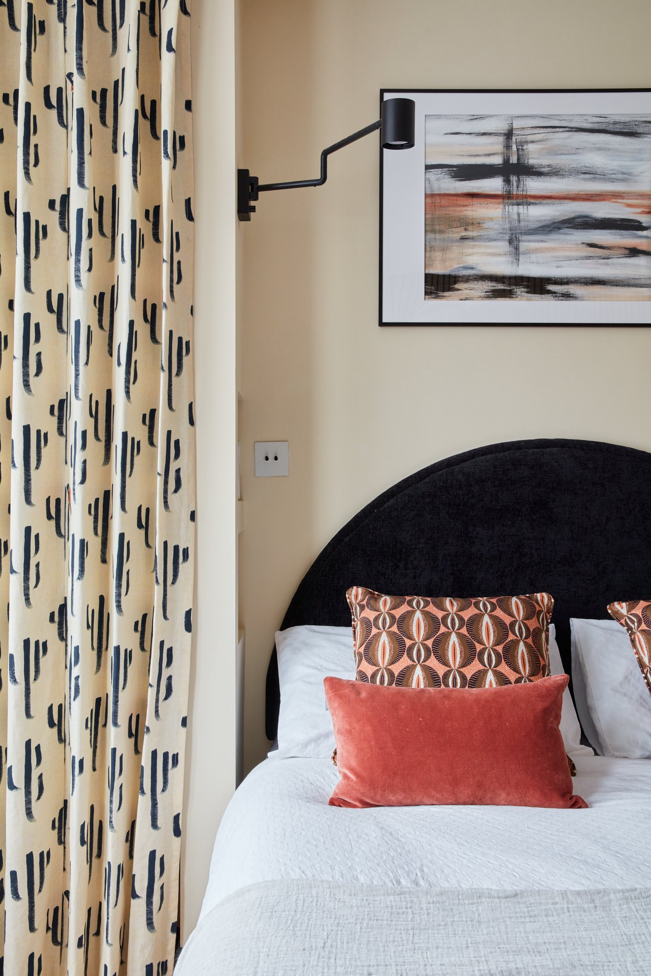 A bedroom with a bed covered in a white quilt, topped with decorative pillows in rust and geometric patterns. A black velvet headboard, a beige lamp with a black arm mounted on the wall, a framed abstract painting, and yellow patterned curtains are v