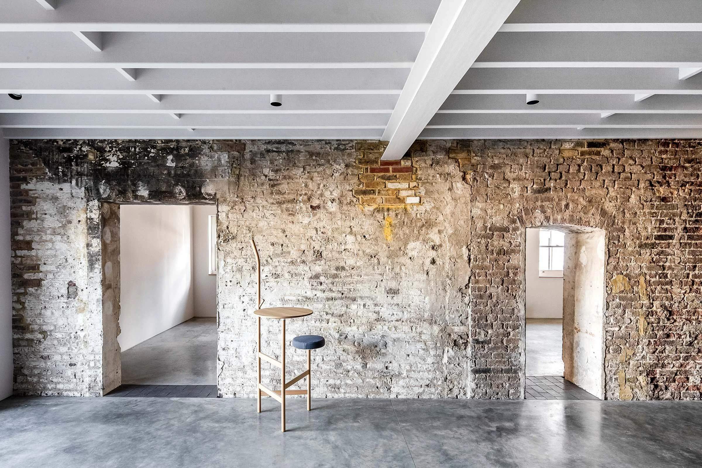 Interior of an unfinished or renovated building with exposed brick walls, a modern white ceiling with beams, a wooden chair with a black cushion, and two doorways leading to rooms with windows.