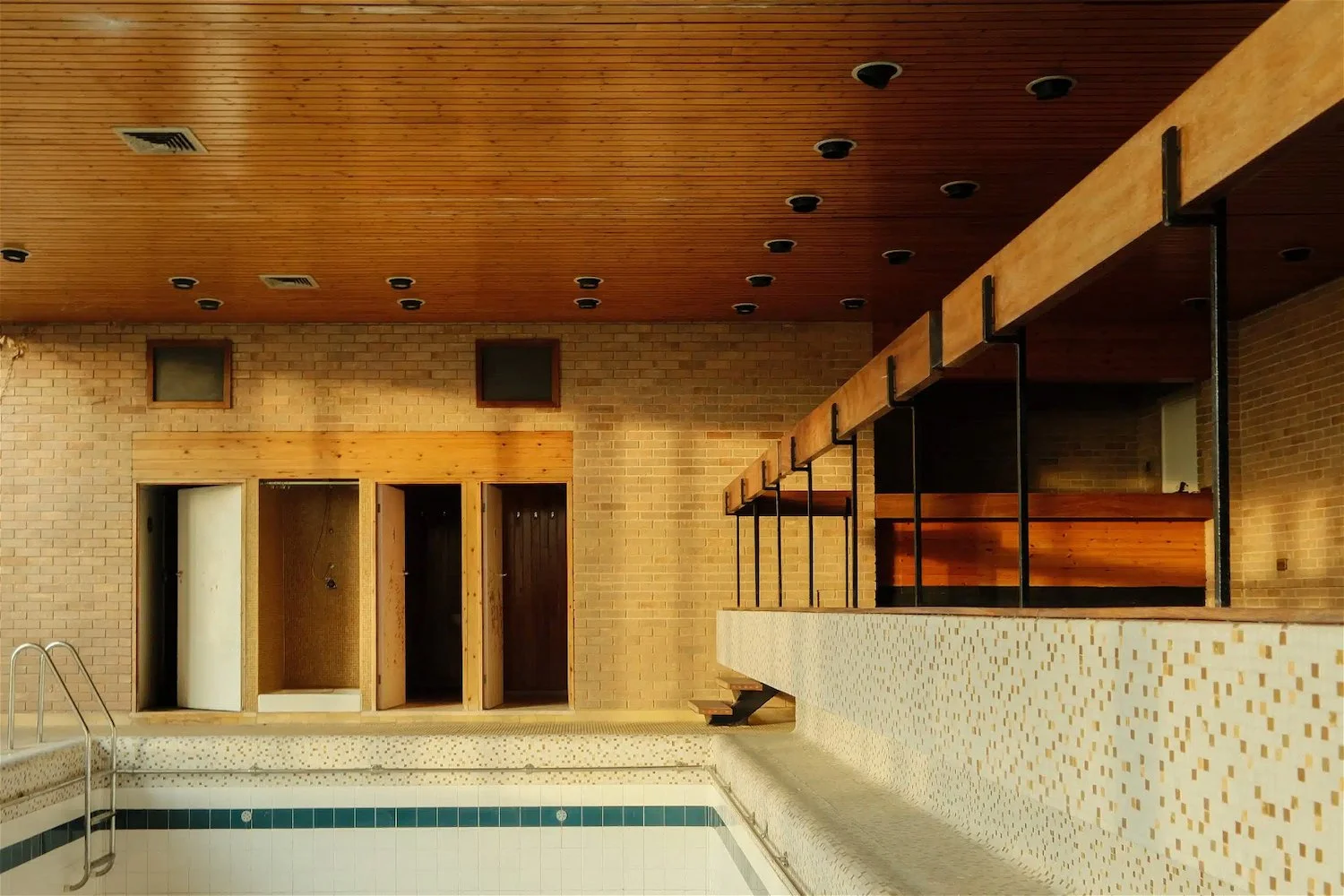 Indoor swimming pool area with brick walls, wooden ceiling, and multiple small windows. The pool has a metal ladder and surrounding mosaic tile edging.
