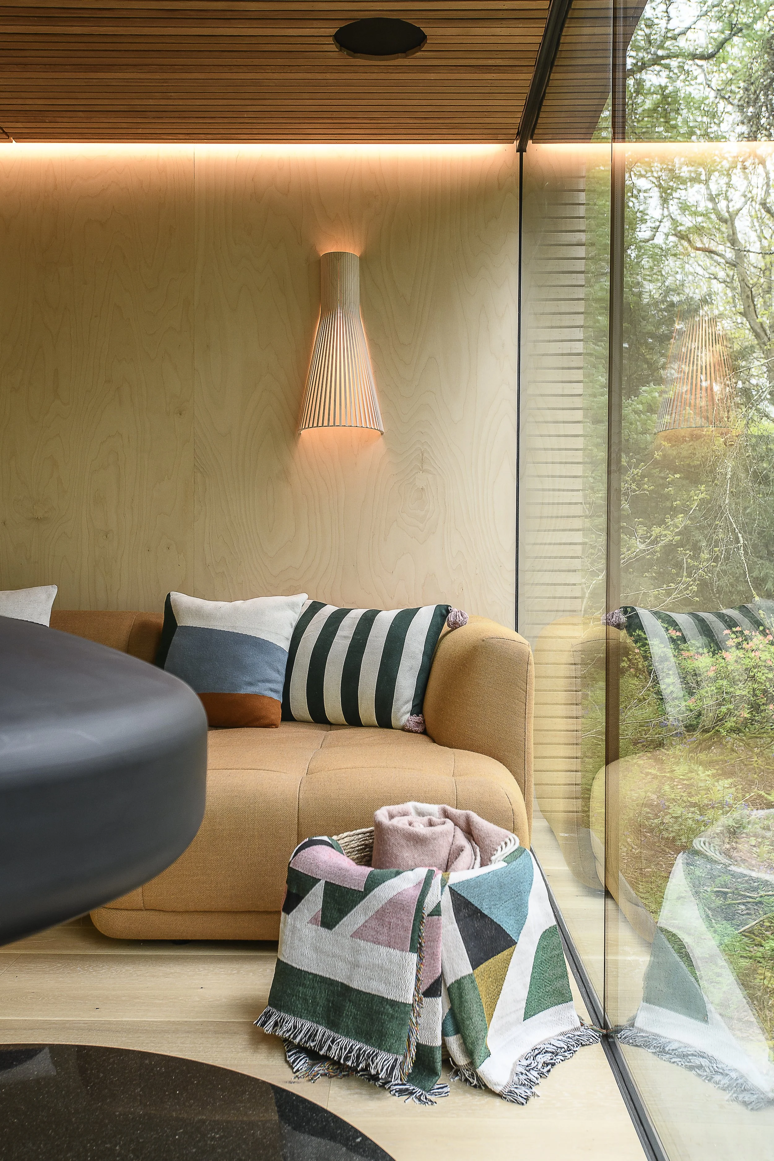 Interior view of a modern living room with a beige sofa, multicolored throw pillows, a wooden wall with a decorative wall sconce, and a large glass window showing outdoor greenery.