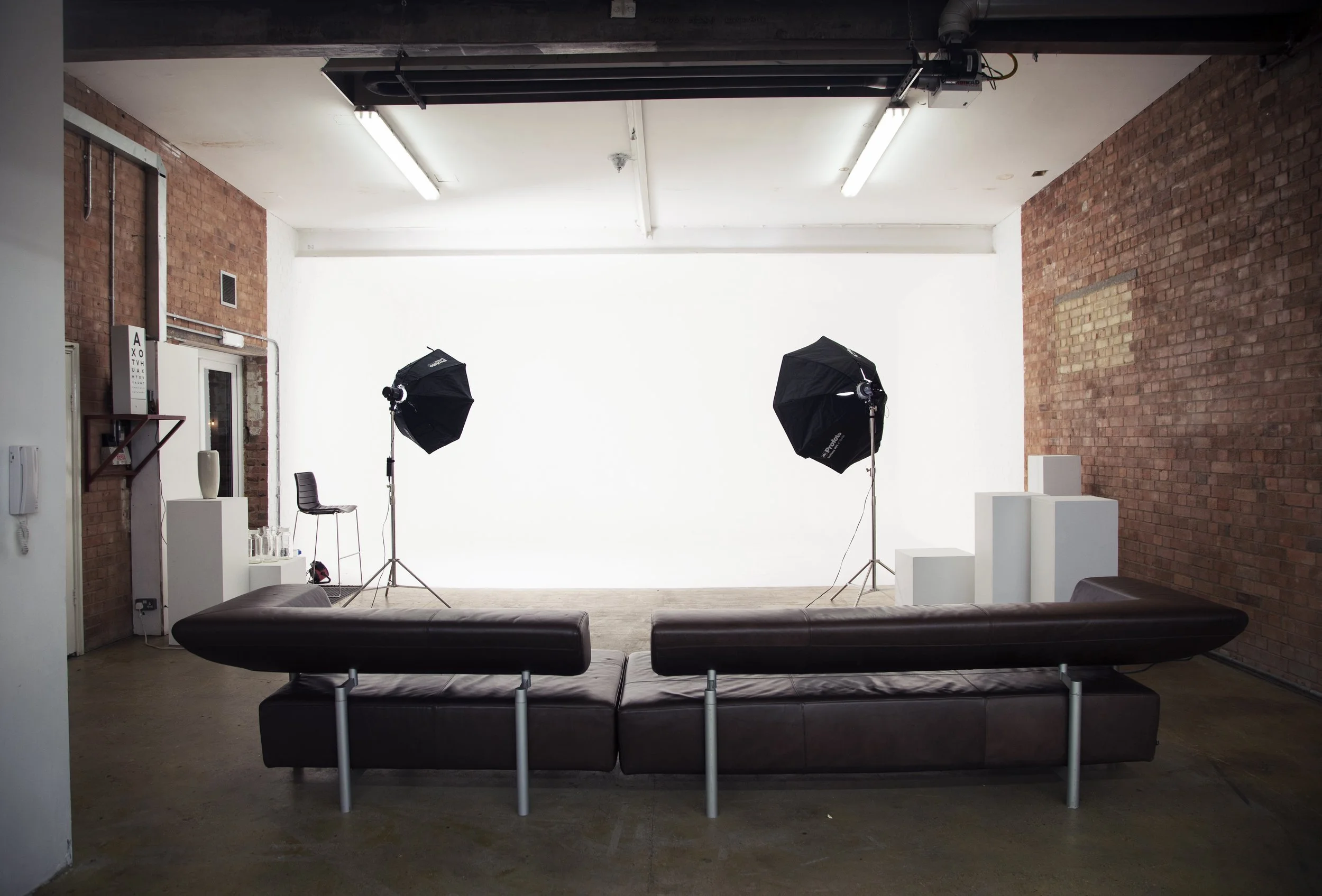 Photography studio with a white backdrop, two studio umbrellas, a black chair, a brown leather sofa, and white pedestals in a brick-walled space.