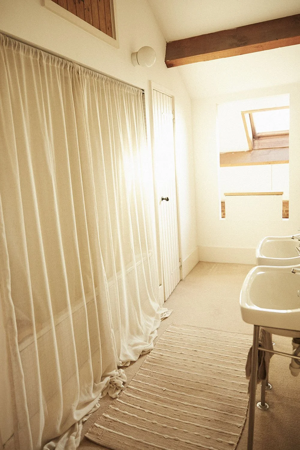 Bright bathroom with beige carpet, white sinks, cream curtains, and a skylight in the ceiling.