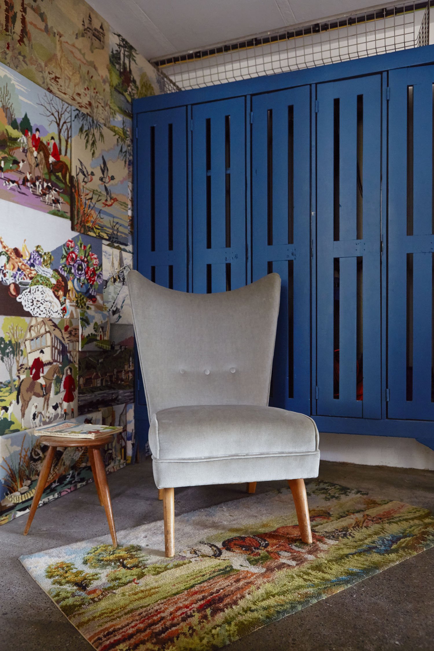 A beige upholstered armchair with wooden legs is placed on a colorful woven rug featuring trees and abstract shapes. To the left of the chair, a small wooden side table holds some magazines. The background includes a wall decorated with vibrant, deta