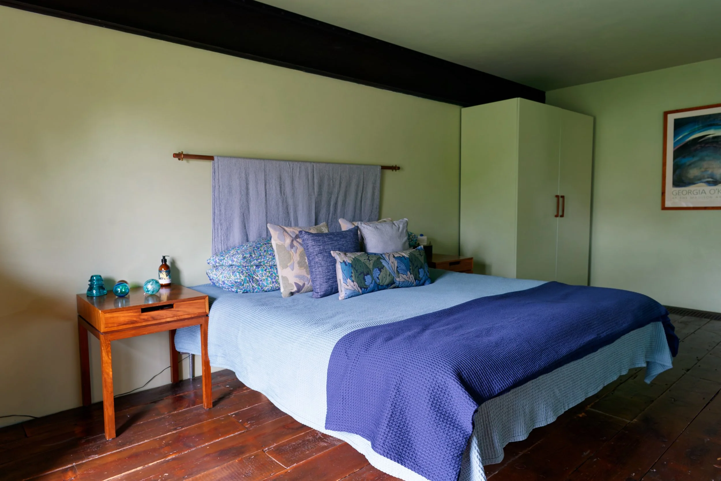 A bedroom with a bed featuring a light blue bedspread and several decorative pillows, a wooden nightstand with small decorative items and a bottle, a wardrobe in beige, and a framed picture on the wall.