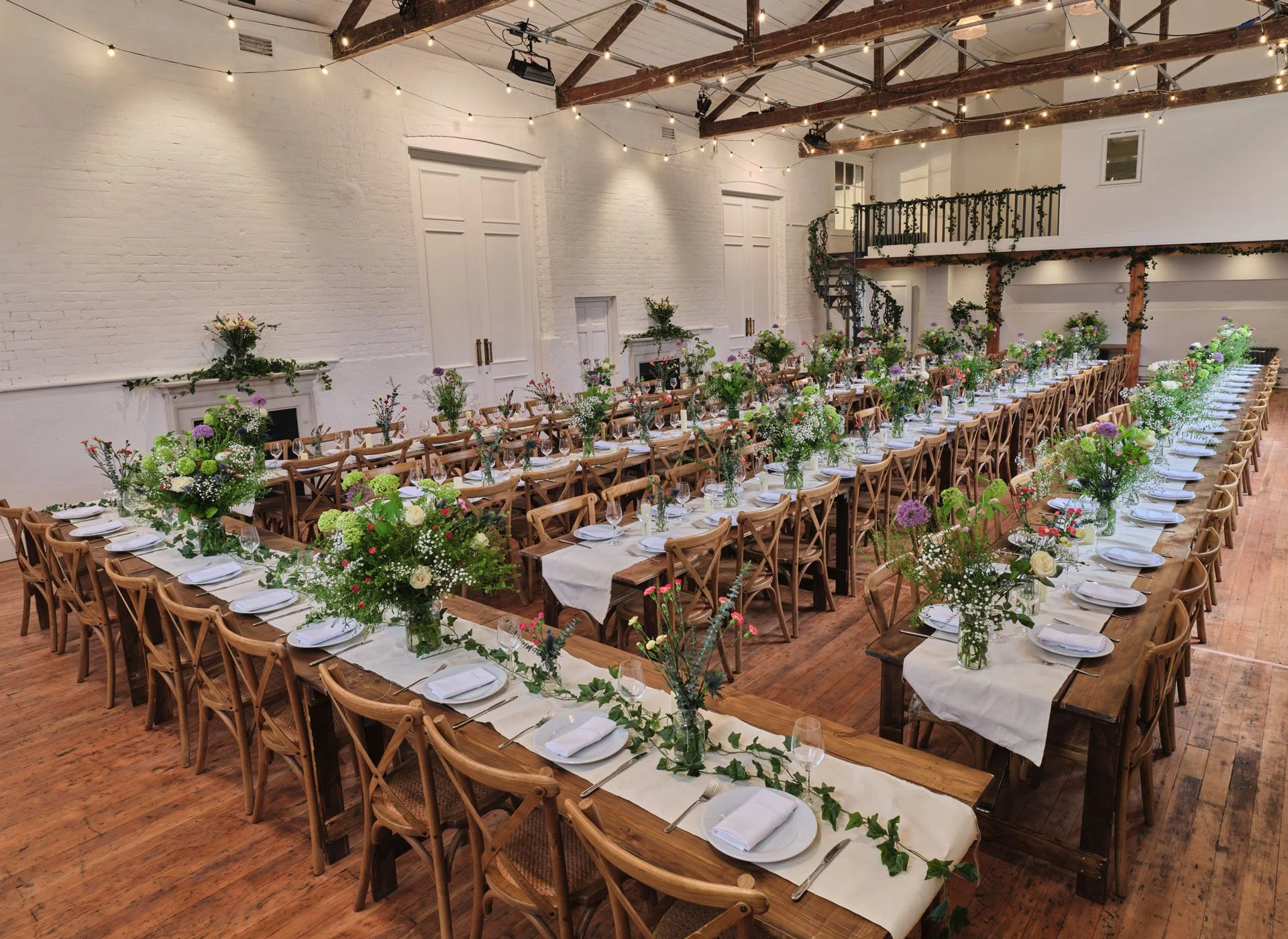 A large indoor banquet hall decorated for a wedding or event with long wooden tables arranged in parallel rows, adorned with white table runners, white plates, silverware, wine glasses, and colorful floral centerpieces in glass vases. The room featur