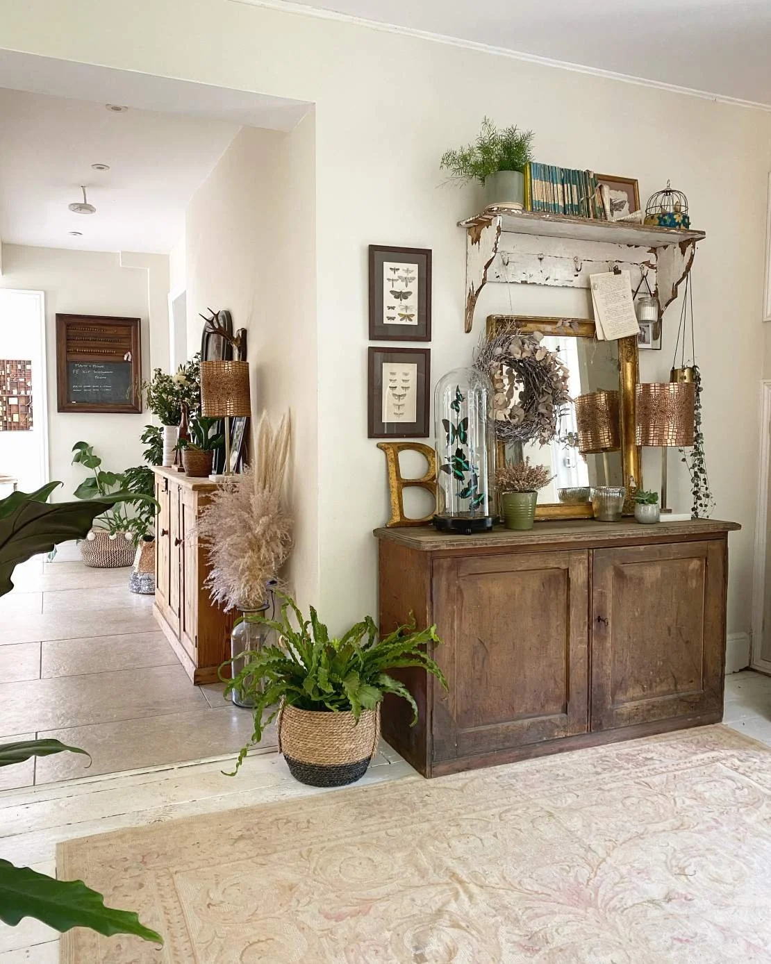 A wooden sideboard with decorative items like plants, framed pictures, and a mirror, positioned against a white wall in a cozy interior space.