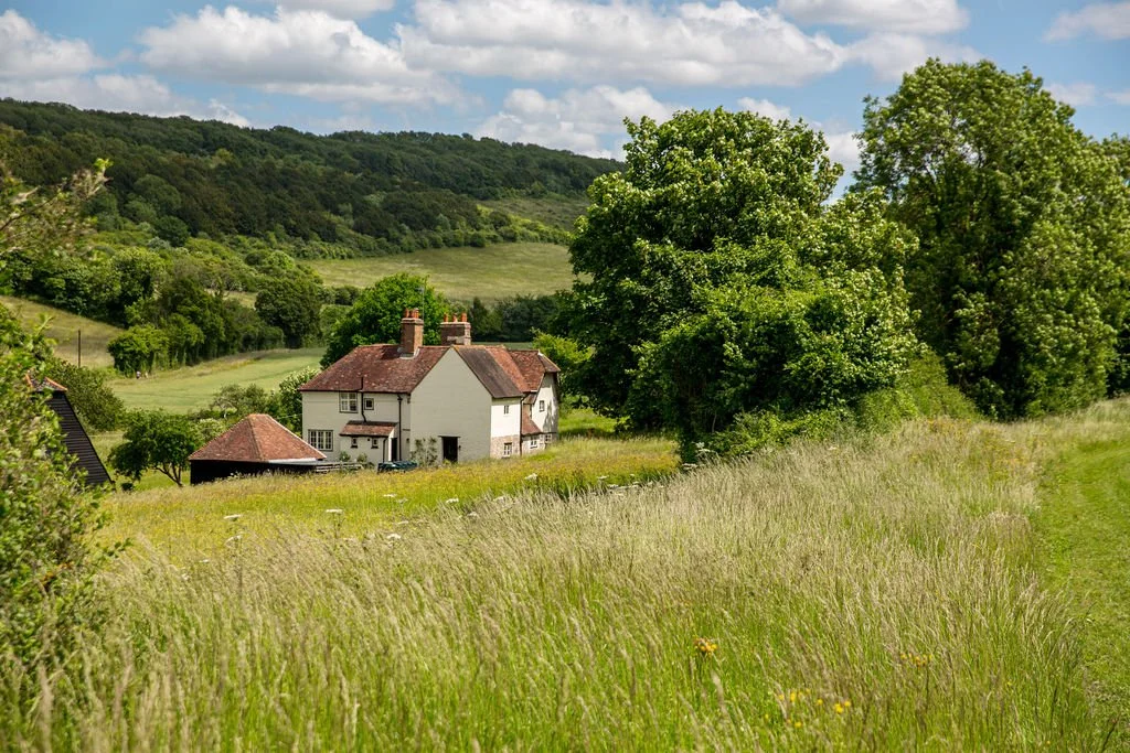 A house with a red roof in a green field with large trees and rolling hills in the background under a partly cloudy sky.