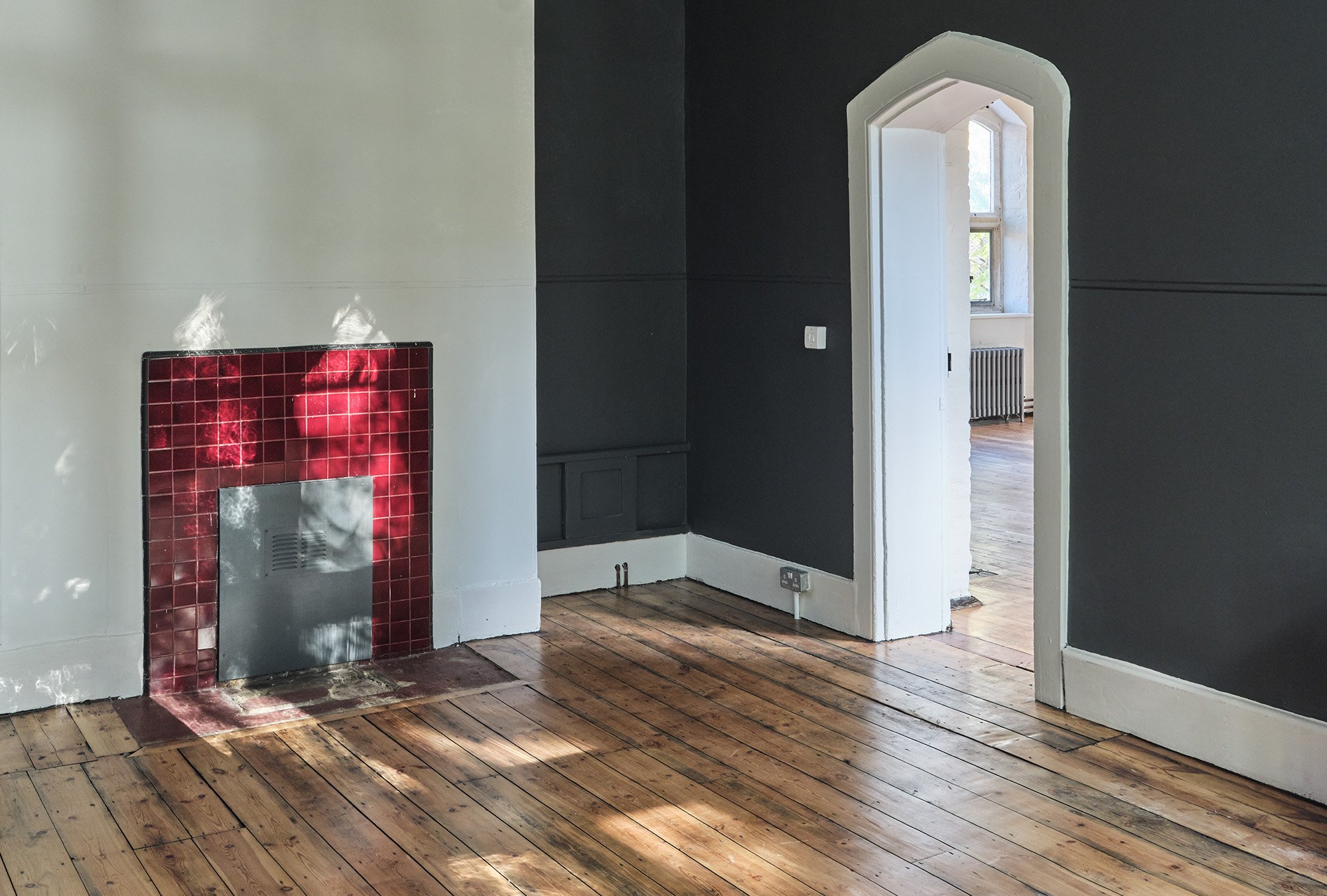 Empty room with wood flooring, white and dark gray walls, a decorative red-tiled fireplace, and a doorway leading to a bright room with a window and radiator.