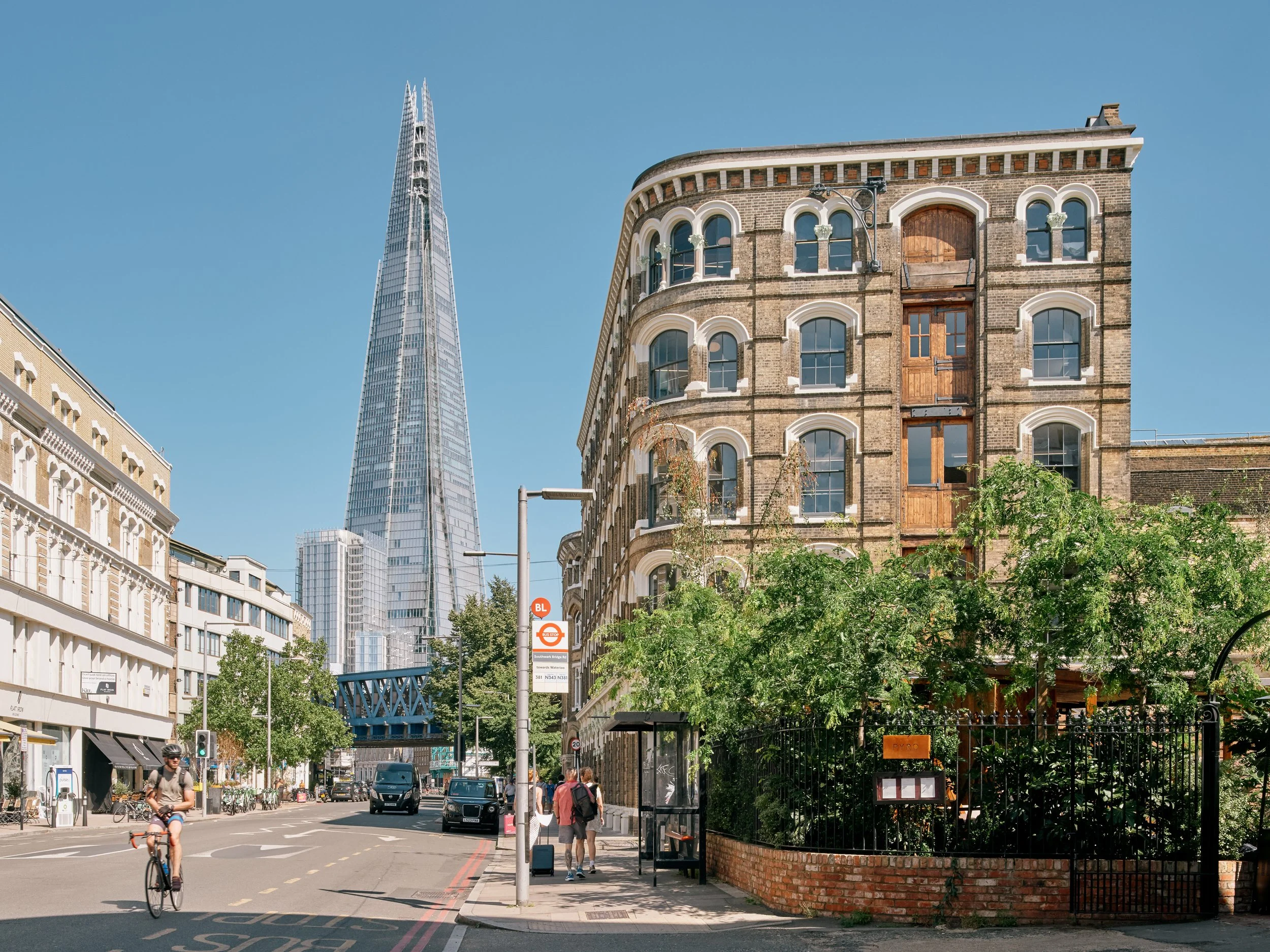 A city street scene with a modern skyscraper, The Shard, in the background, and older brick buildings, trees, pedestrians, and a cyclist in the foreground.