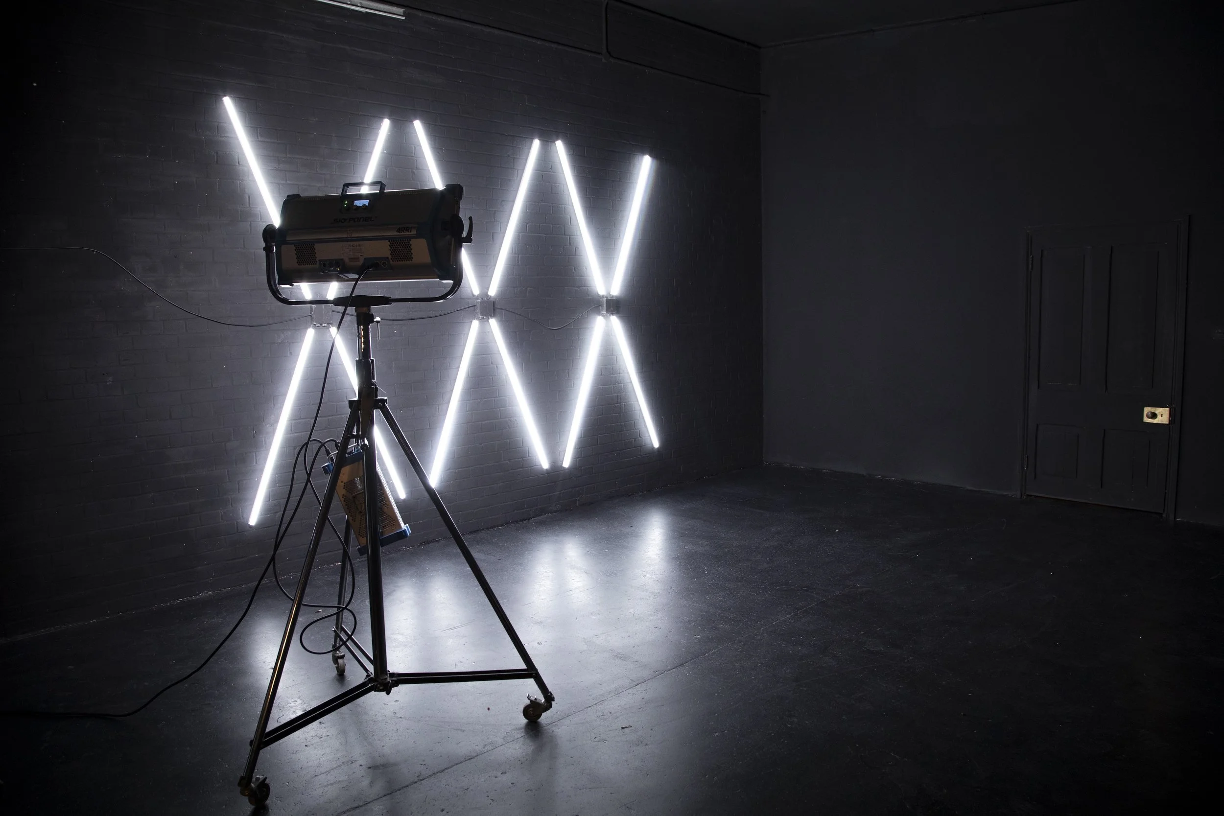 Empty room with black walls and floor, illuminated by a grid of white vertical and diagonal LED lights on one wall, and a professional video or photography light on a tripod stand in the foreground.