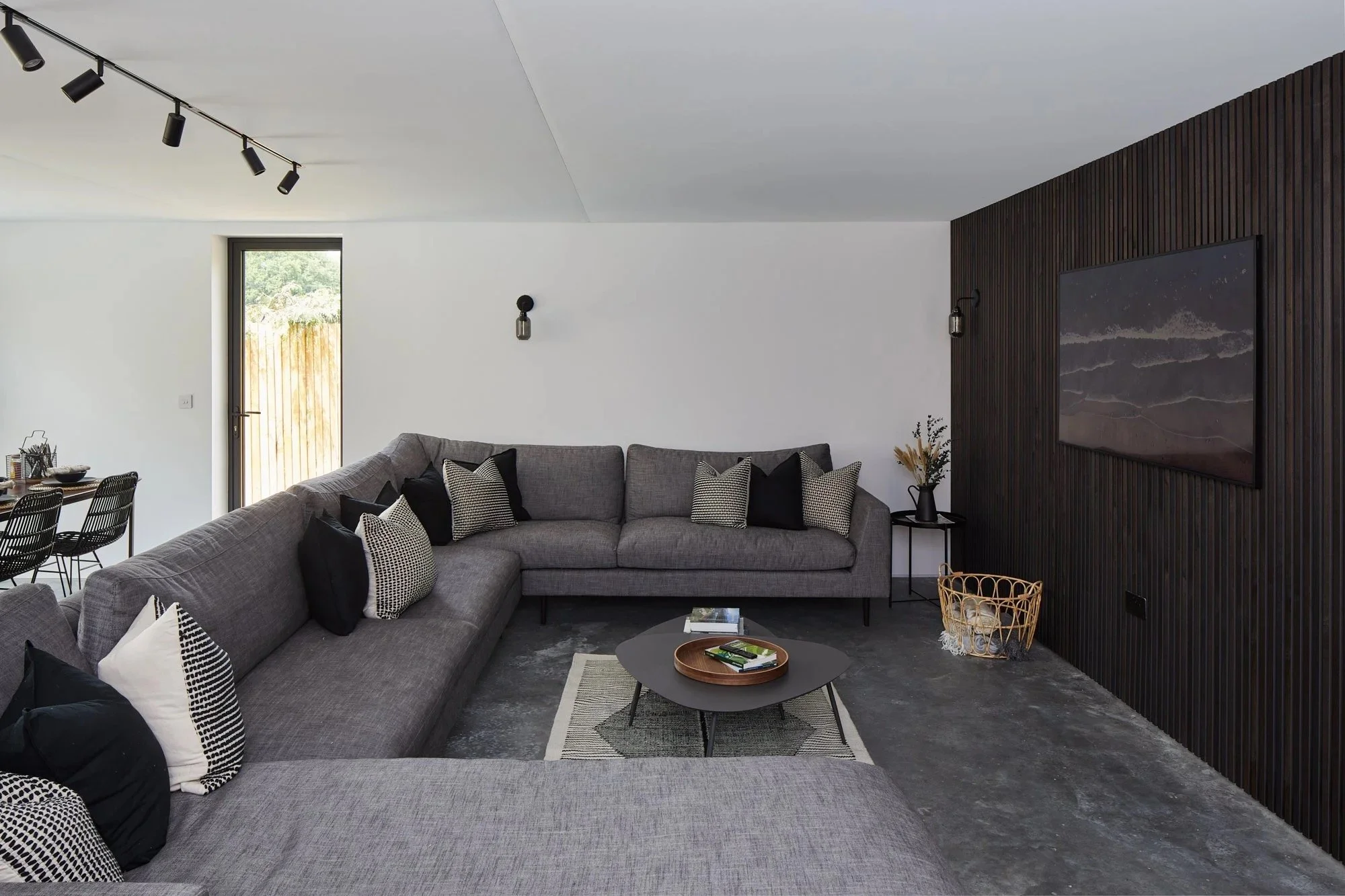 Modern living room with a gray sectional sofa against a white wall, black and patterned throw pillows, a coffee table with books, a dark wood accent wall with a large mounted television, and minimal side tables with decorative items. A doorway with a