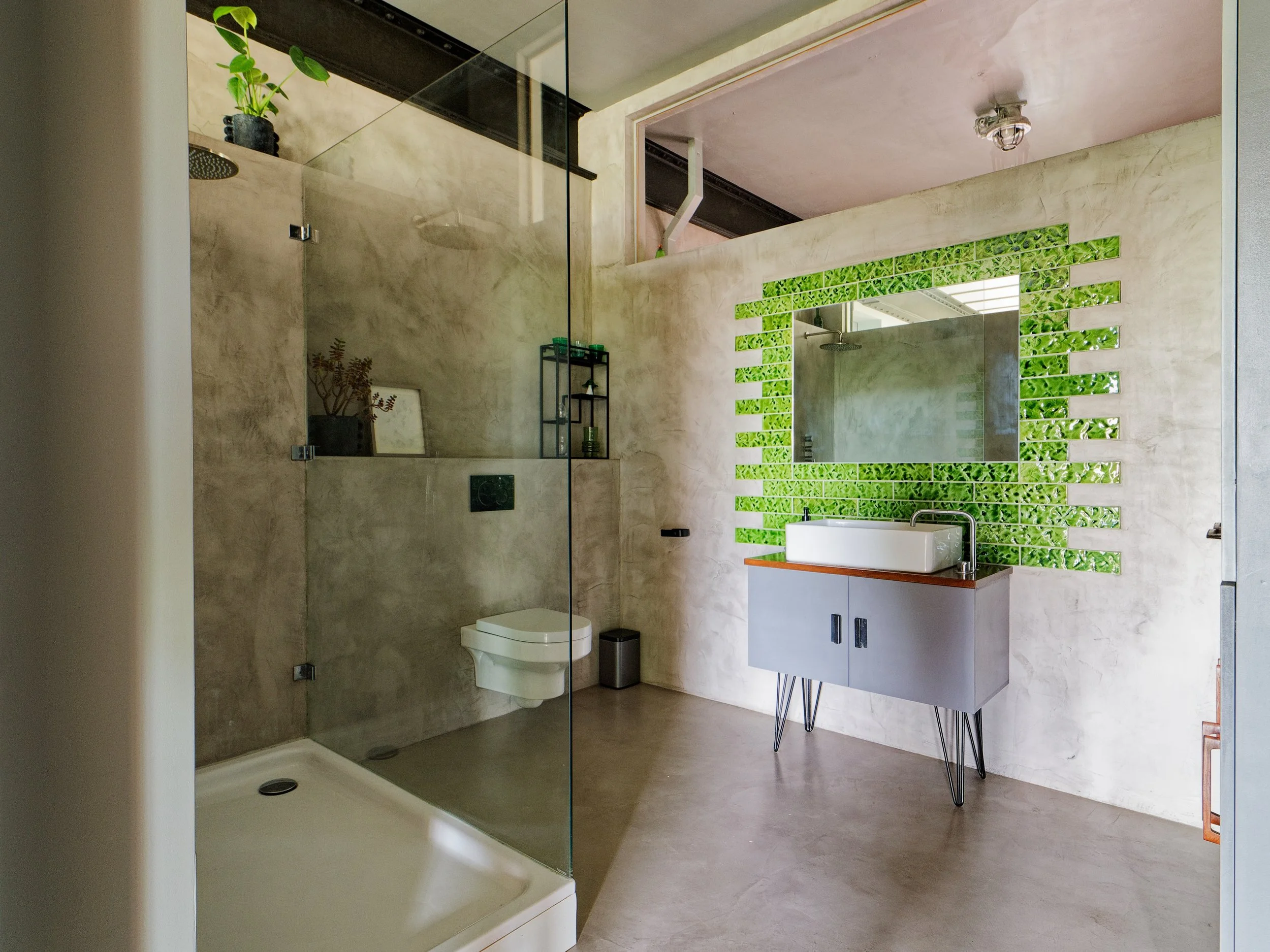 Modern bathroom with concrete-looking walls, a walk-in glass shower, a wall-mounted toilet, and a gray vanity with a white basin sink and a green tiled mirror. There are potted plants on shelves and a mirror reflecting greenery.