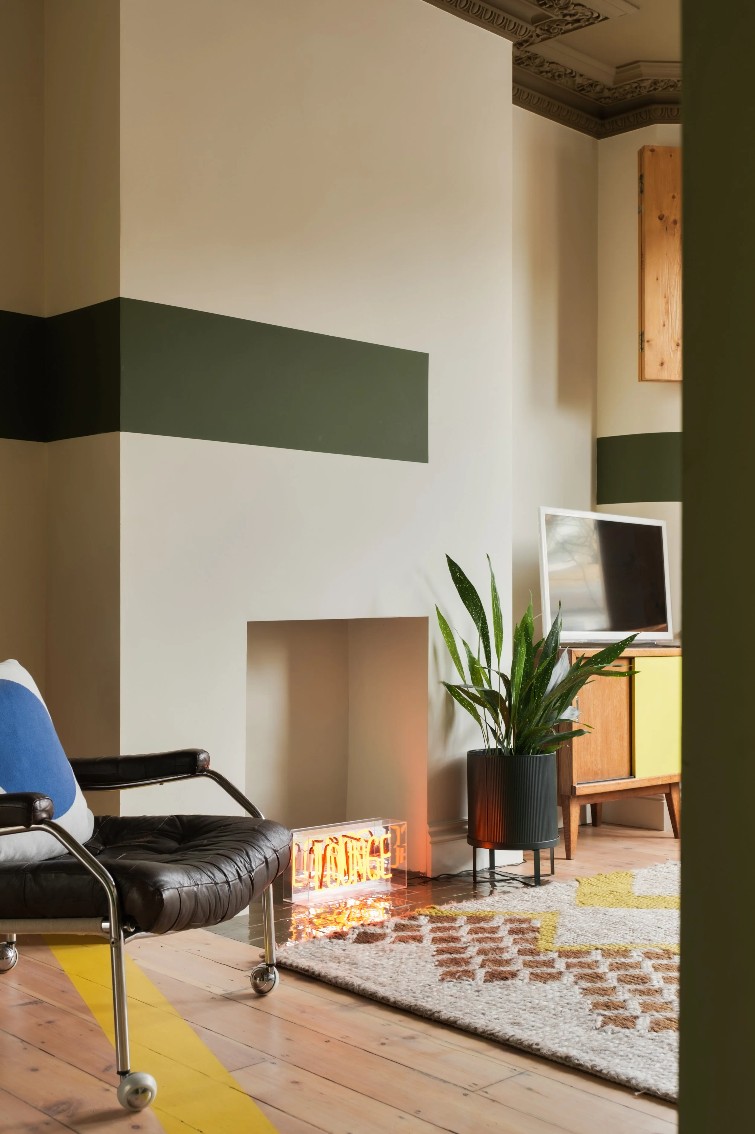 A living room with a black leather armchair, a potted plant, a television on a wooden cabinet, a patterned rug, and a decorative brick fireplace.