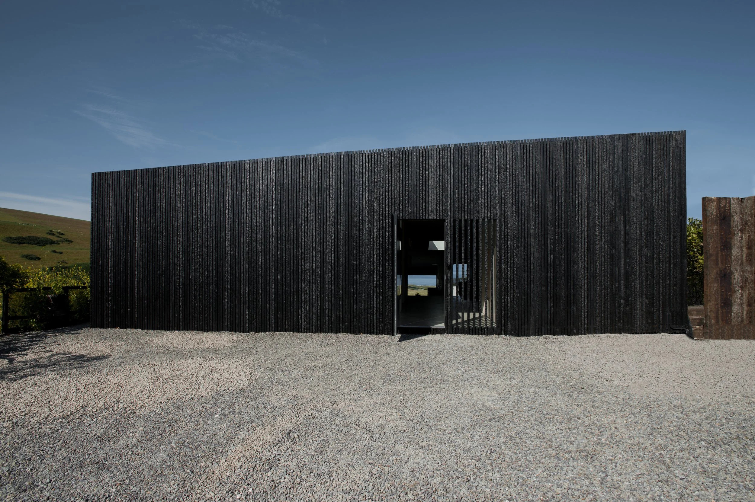 Modern black wooden building with an open doorway, gravel ground, green hillside, and blue sky in the background.