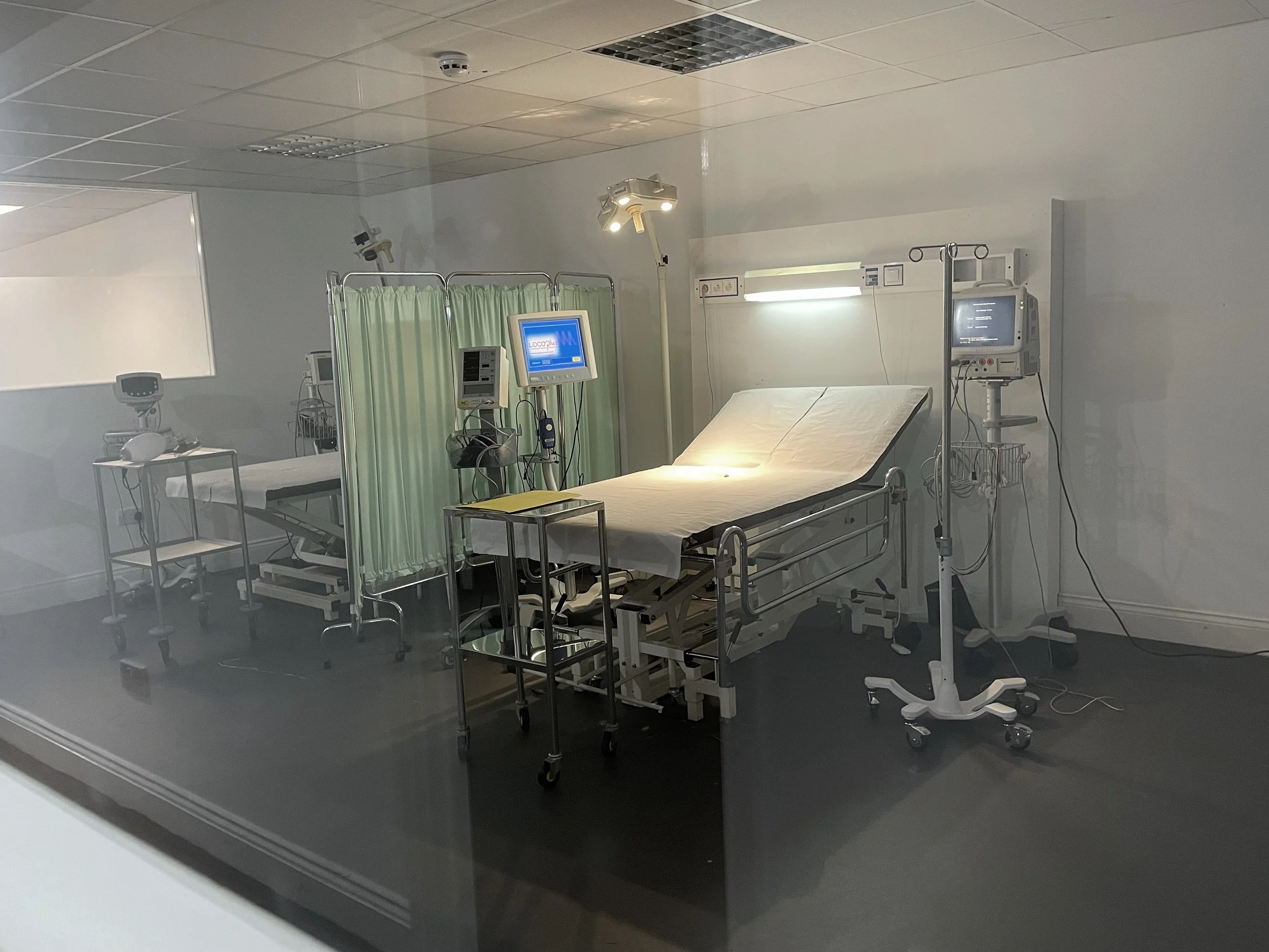 An empty hospital emergency room with a hospital bed, medical monitors, and equipment, separated by a green privacy curtain.