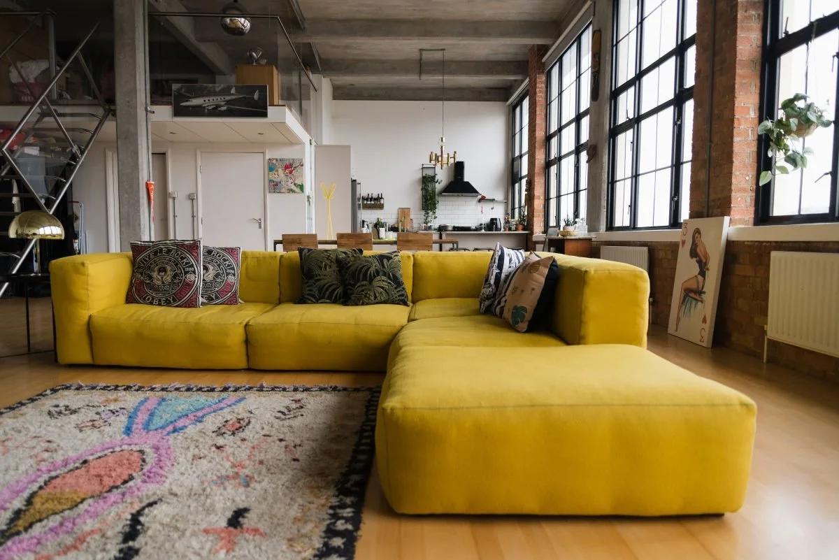A spacious loft-style living room with large windows, a yellow sectional sofa with decorative pillows, a patterned area rug, and an open kitchen and dining area in the background.