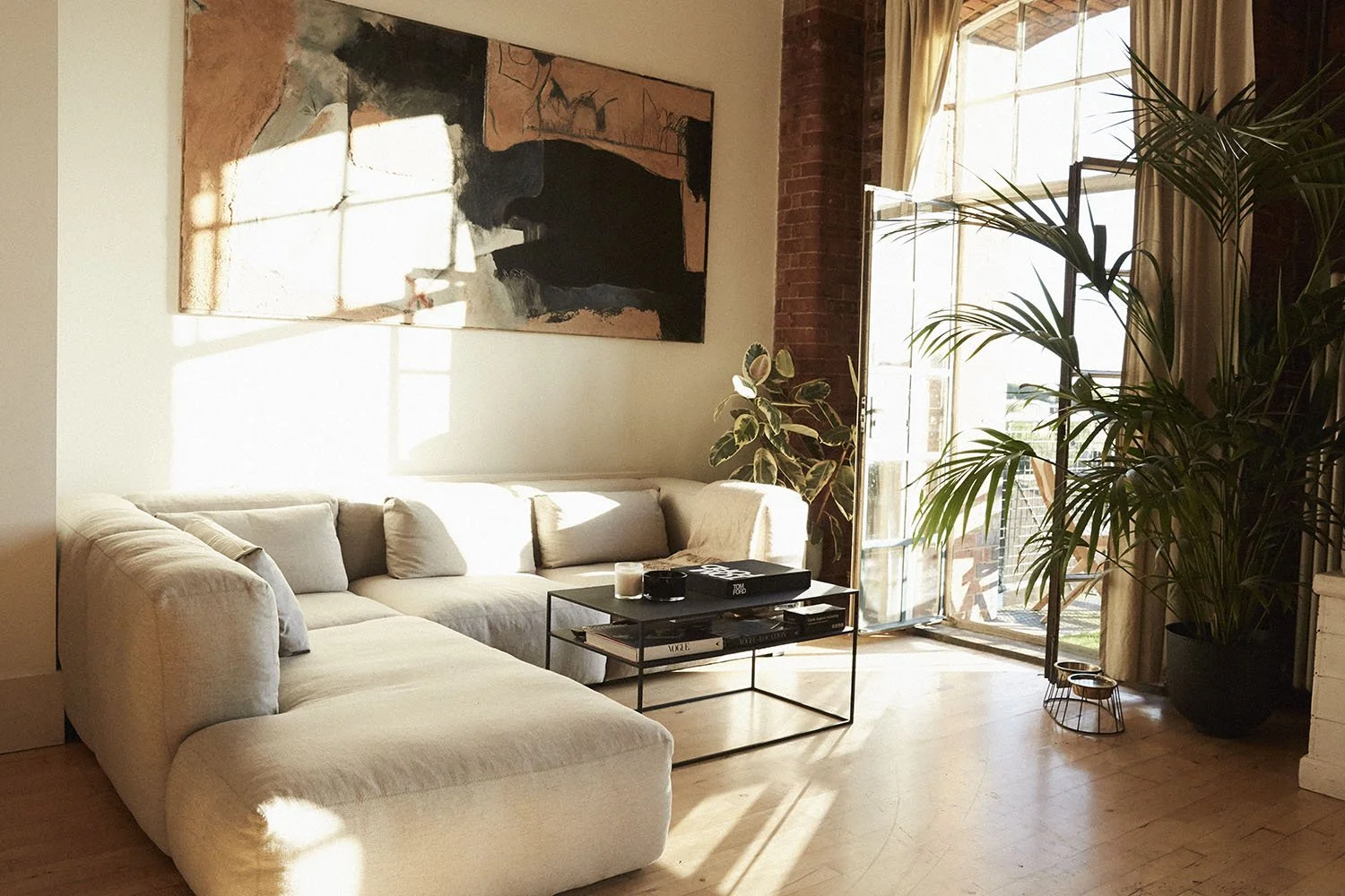 Bright living room with a light-colored sectional sofa, a coffee table with books, candles, and a tray, large potted plants, a mural painting on the wall, and sunlight streaming through open glass doors with beige curtains.