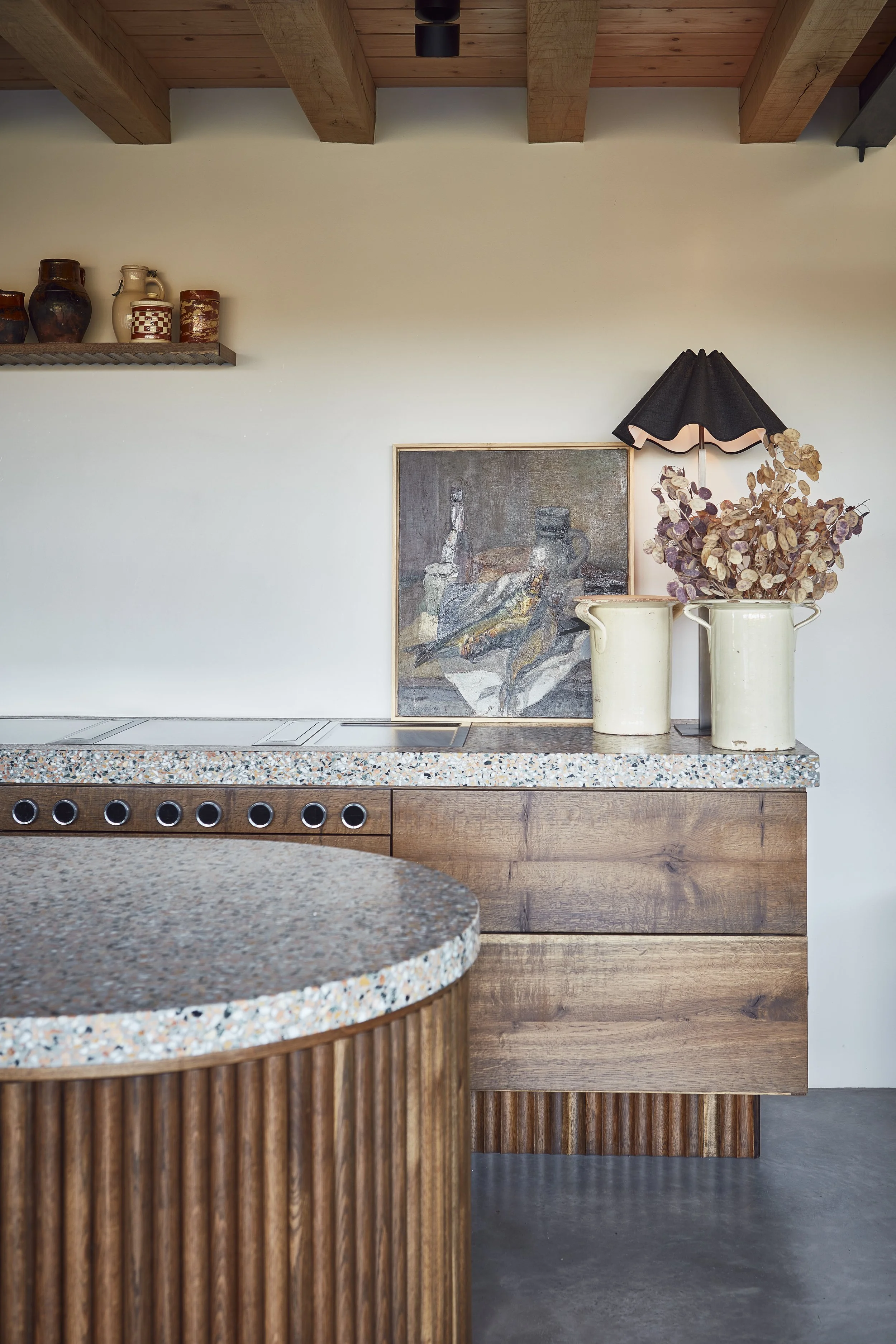 A modern interior space with a wooden ceiling and beige wall. Decor includes a floating shelf with pottery and a framed artwork leaning against the wall. Two light-colored vases containing dried foliage and a black lampshade are on a table.