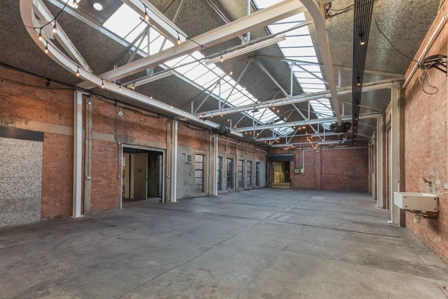 Empty industrial-style warehouse with exposed brick walls, large skylights, string lights, and concrete floor.