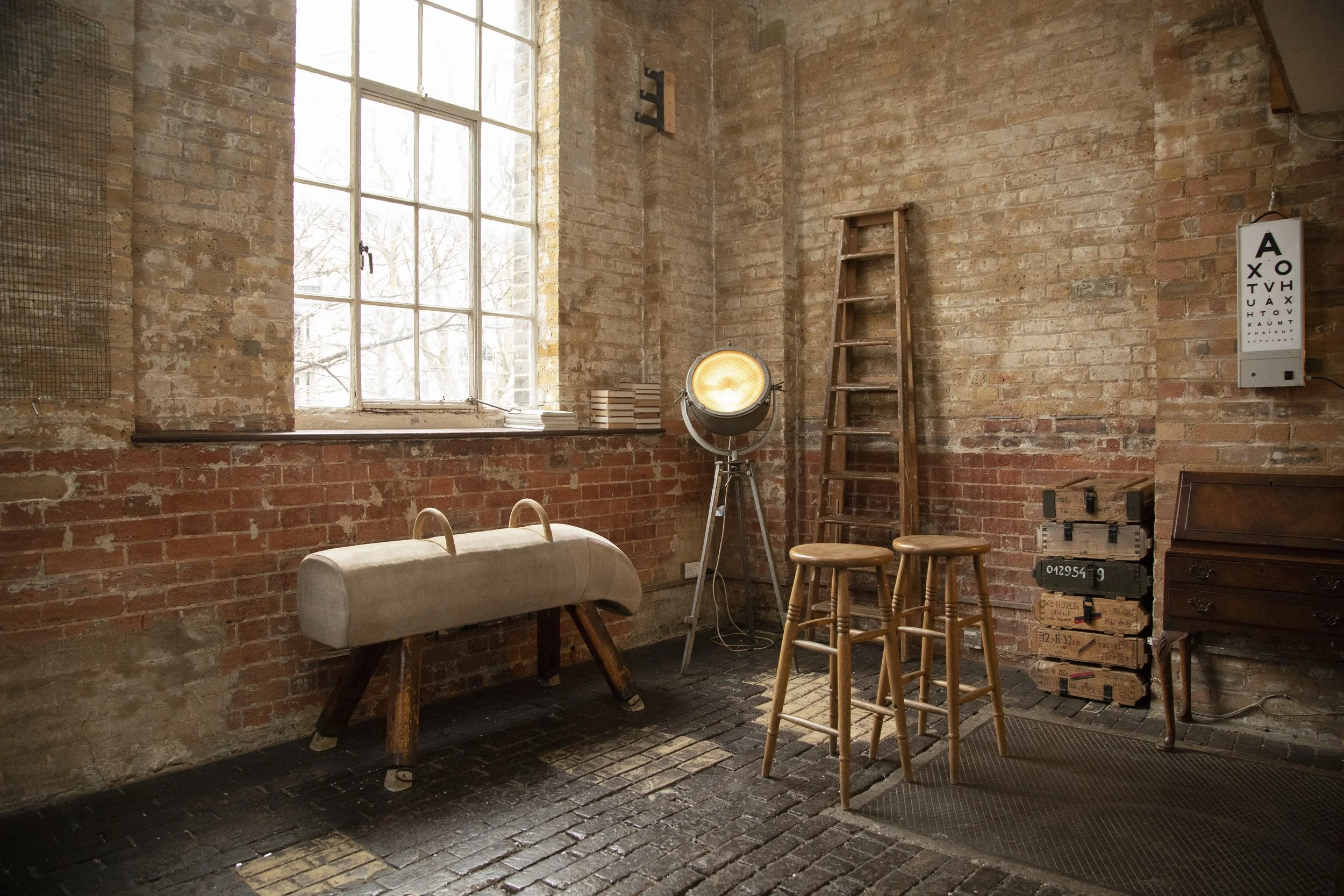 An interior view of a rustic room with exposed brick walls, large window with white framework, a vintage spotlight, a ladder, three wooden stools, a soft gymnastic vault, and a chest of drawers.
