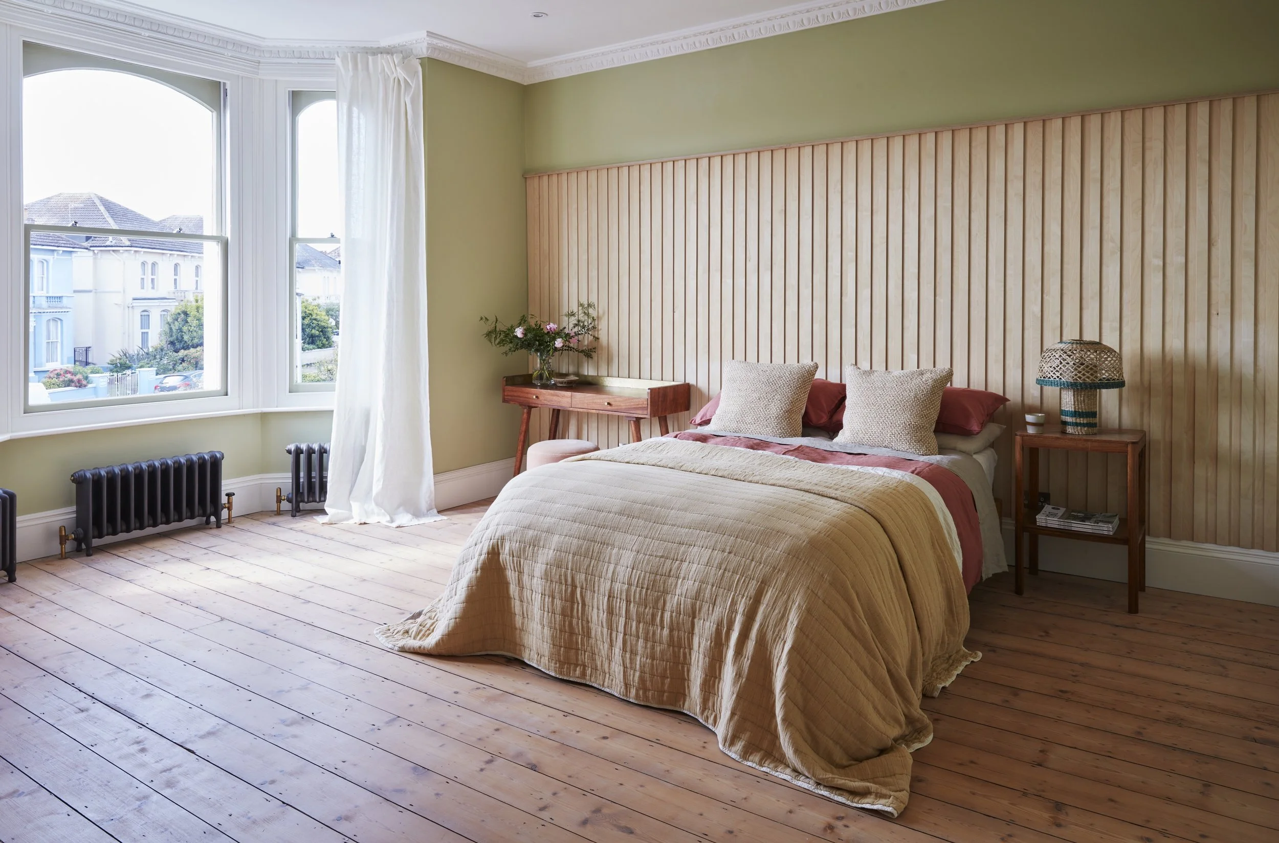 Bright bedroom with a large window, wooden floor, and a bed with a beige quilt and pillows. There are two nightstands with lamps and a flower vase, along with a wooden accent wall behind the bed.
