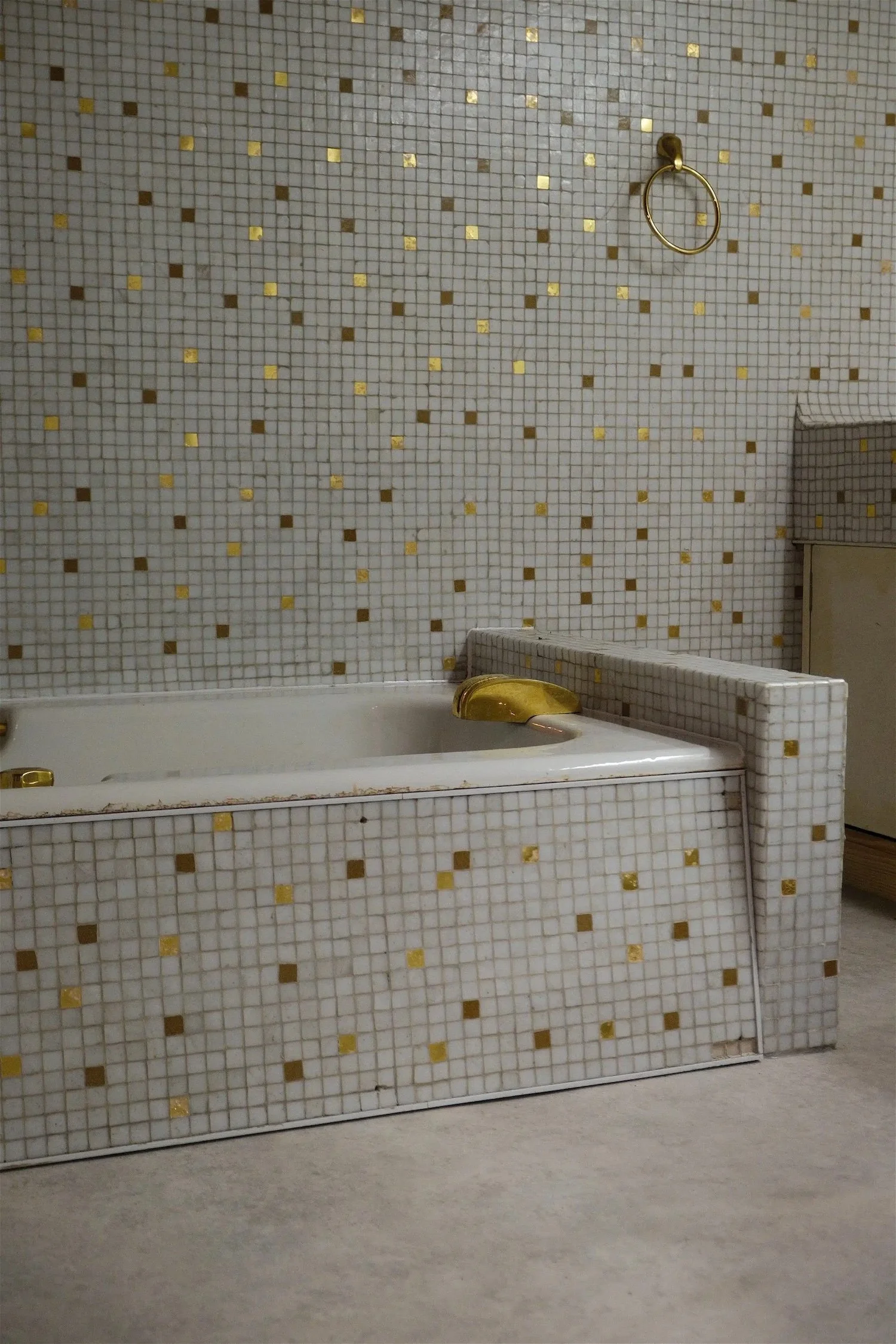 A vintage bathroom with a built-in bathtub covered in small white tiles with gold accents, gold fixtures, and a wall-mounted towel ring.