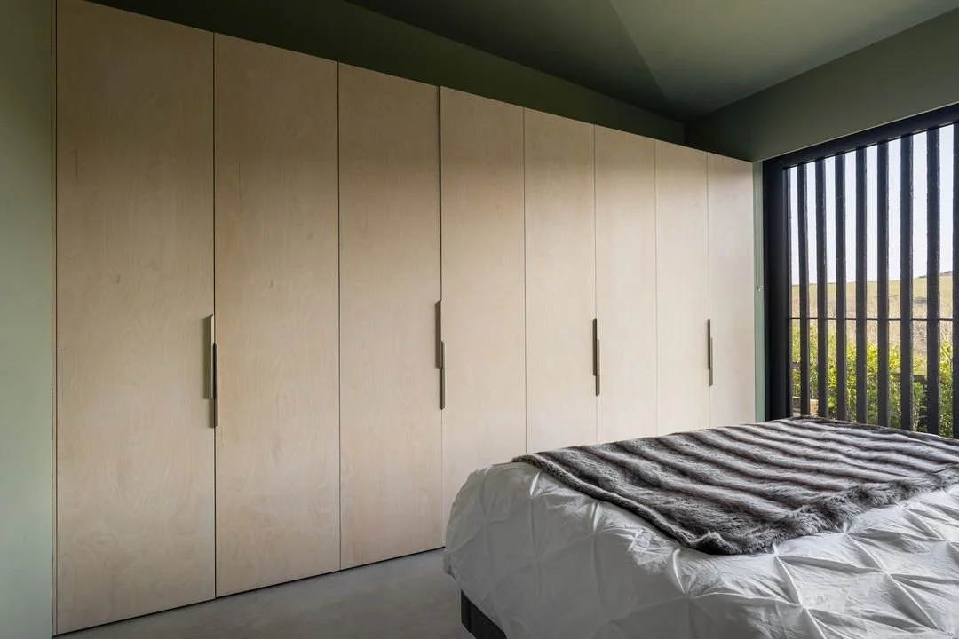 View of a bedroom with a large built-in wardrobe with light wood doors, a bed with white bedding, and a window with black vertical bars overlooking greenery.