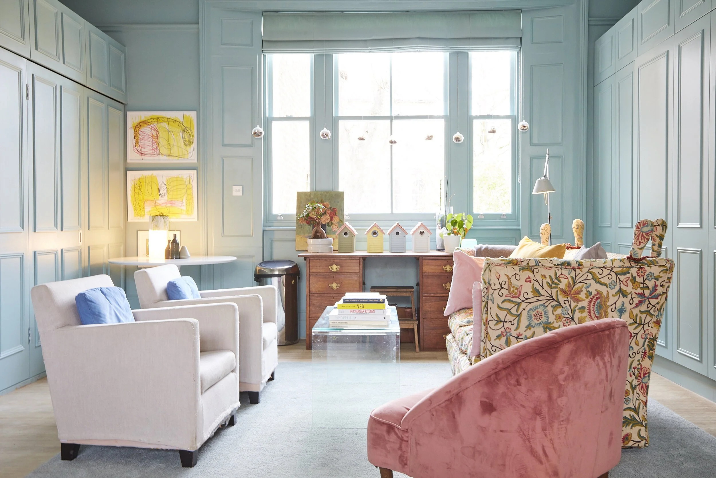 Colorful living room with light blue paneled walls, large windows, white and patterned chairs, a pink velvet armchair, a wooden sideboard with birdhouse decorations, and wall art with children's drawings.