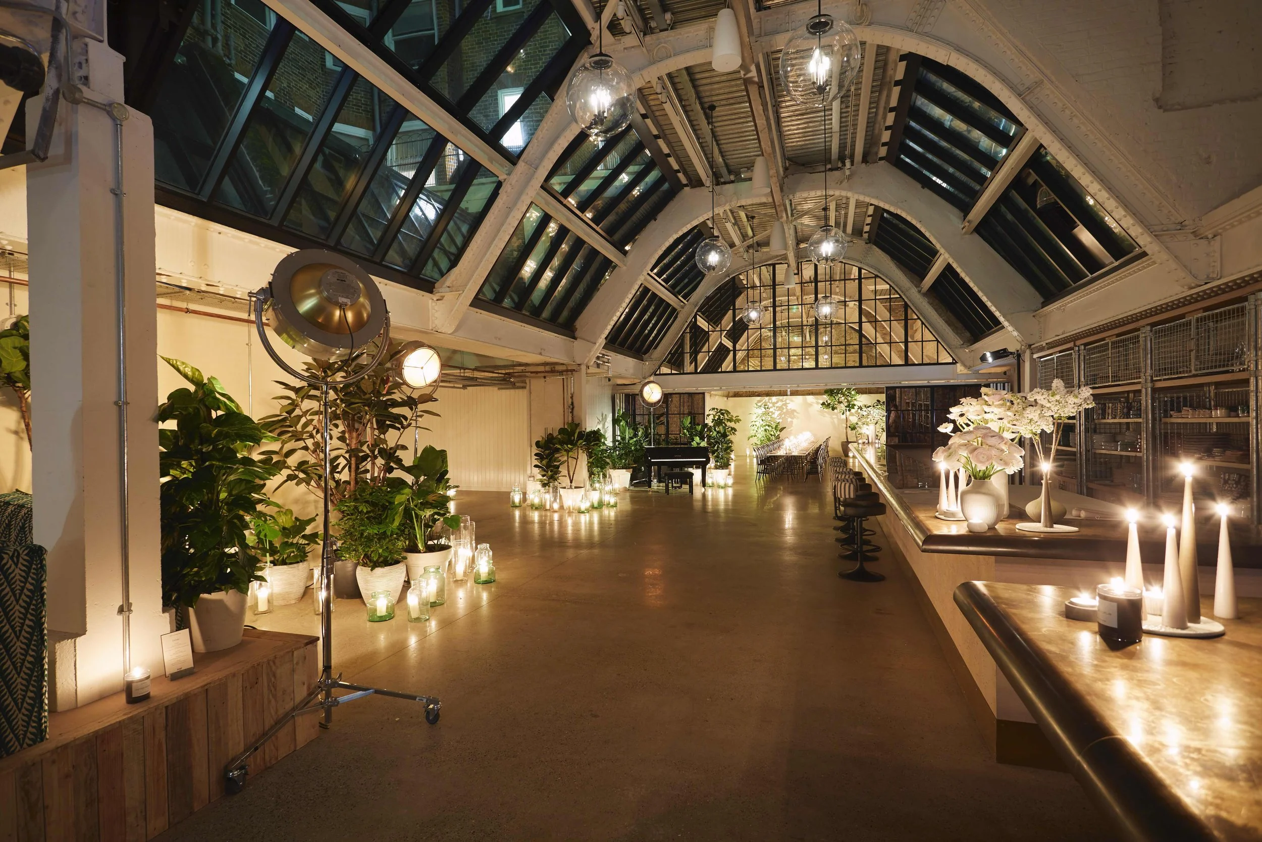 Elegant indoor space with high arched glass ceiling, decorated with plants and flowers, candles, and soft lighting, featuring a piano at the far end.