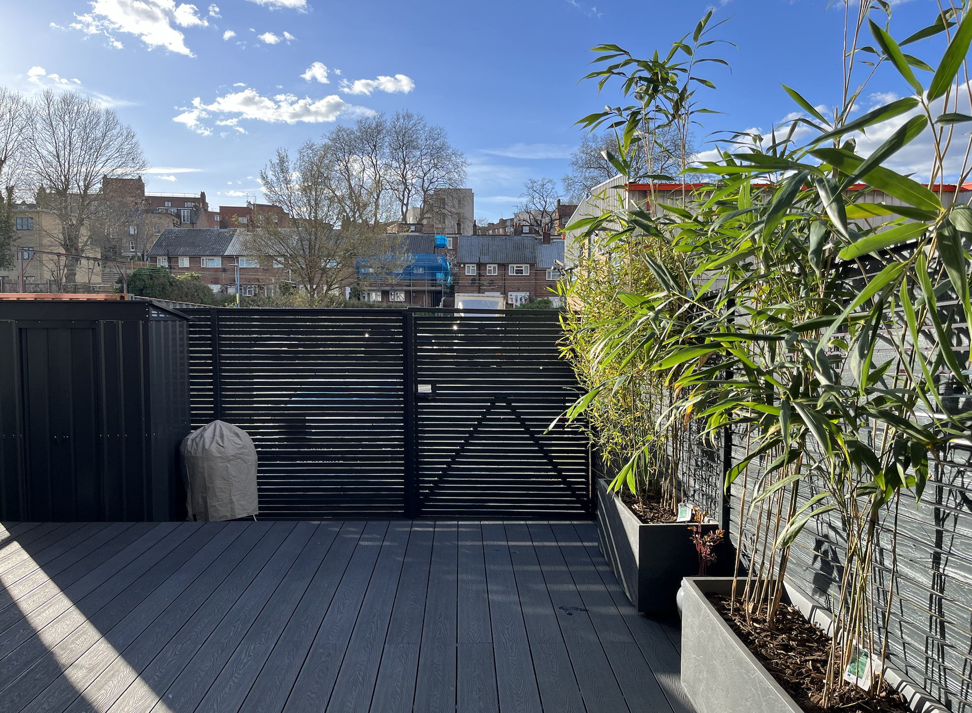 A modern outdoor balcony with black wooden flooring and fencing, large potted bamboo plants on a sunny day with a blue sky and scattered clouds, residential buildings in the background.