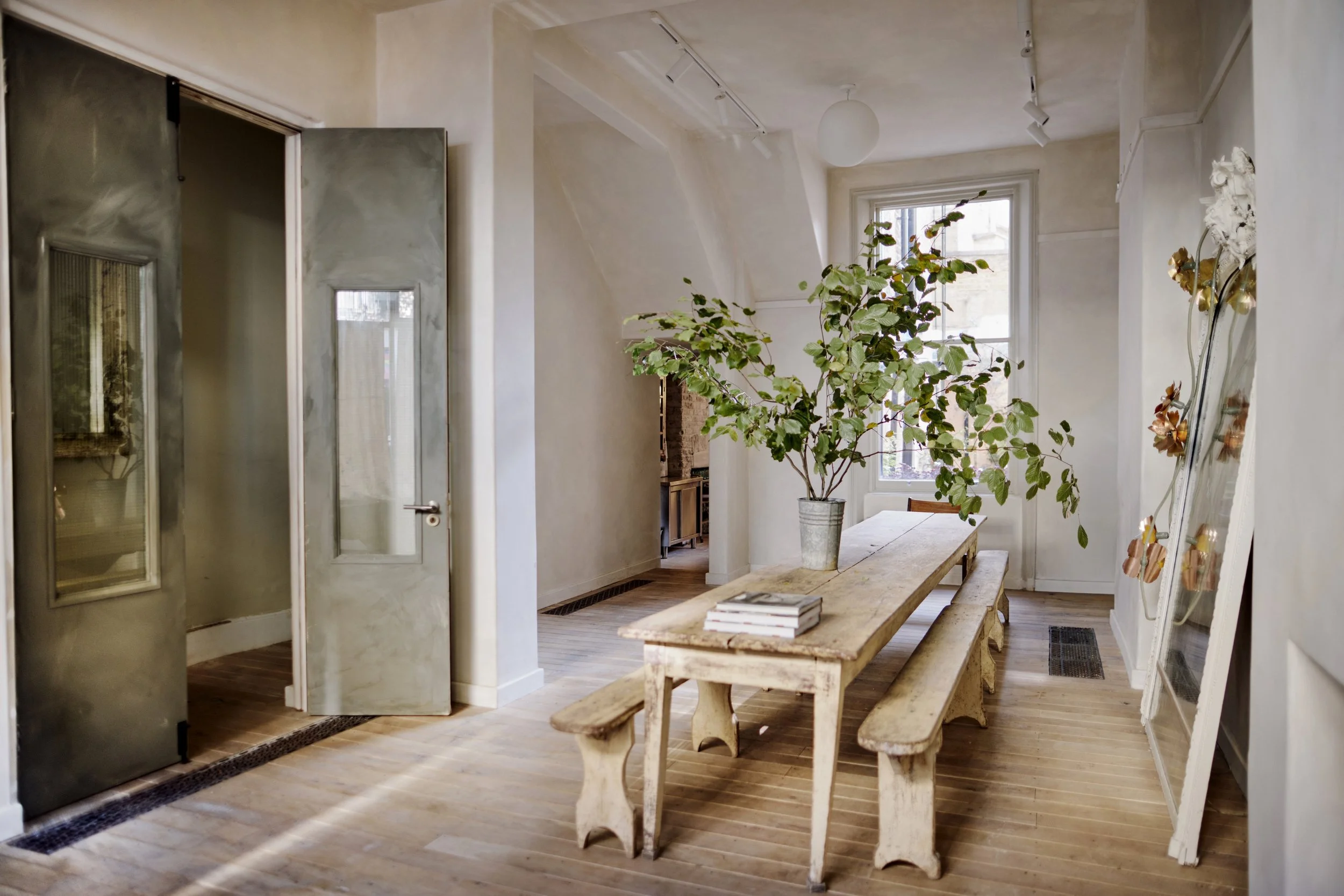 A cozy dining room with wooden floors, a rustic wooden table, matching benches, a large potted plant, and a window letting in natural light.