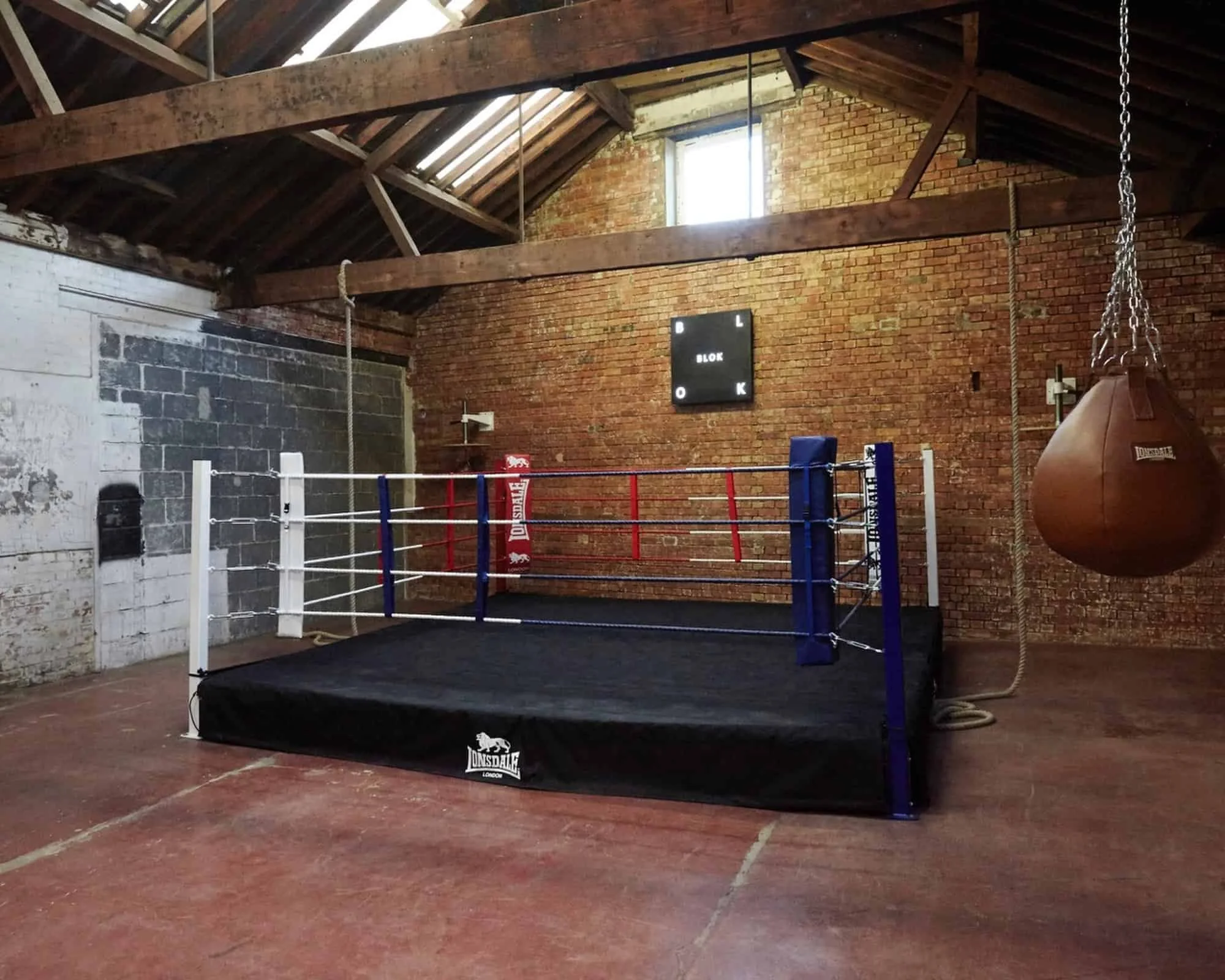Boxing ring with red, white, and blue ropes in an industrial-style gym with exposed brick walls, wooden ceiling beams, a punching bag, and a black clock on the wall.