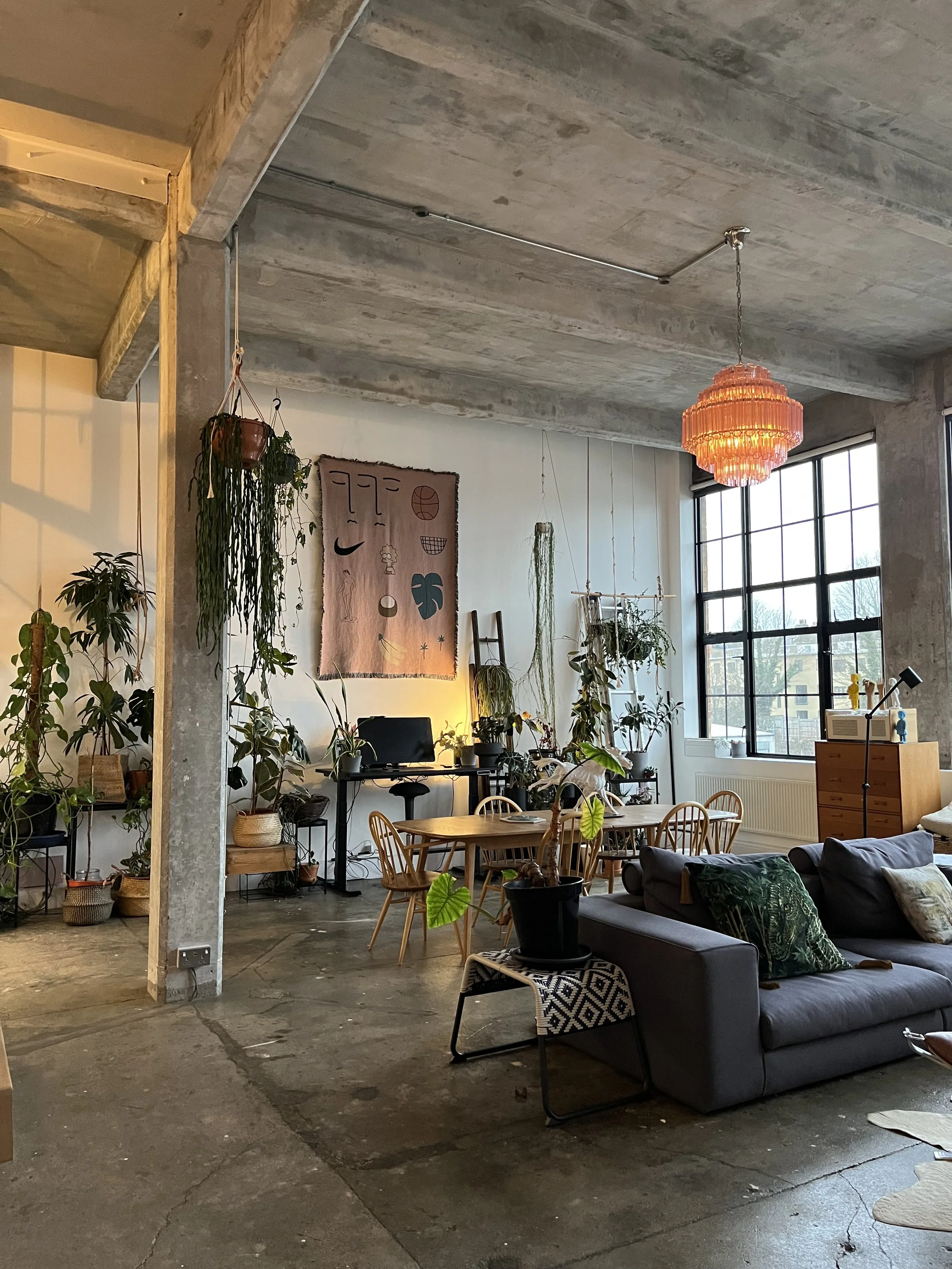 Industrial-style living room with large windows, concrete ceiling, and various plants, including a hanging planter and potted plants on tables and shelves. A black desk with a computer, a wooden dining table with chairs, a gray sofa with decorative p