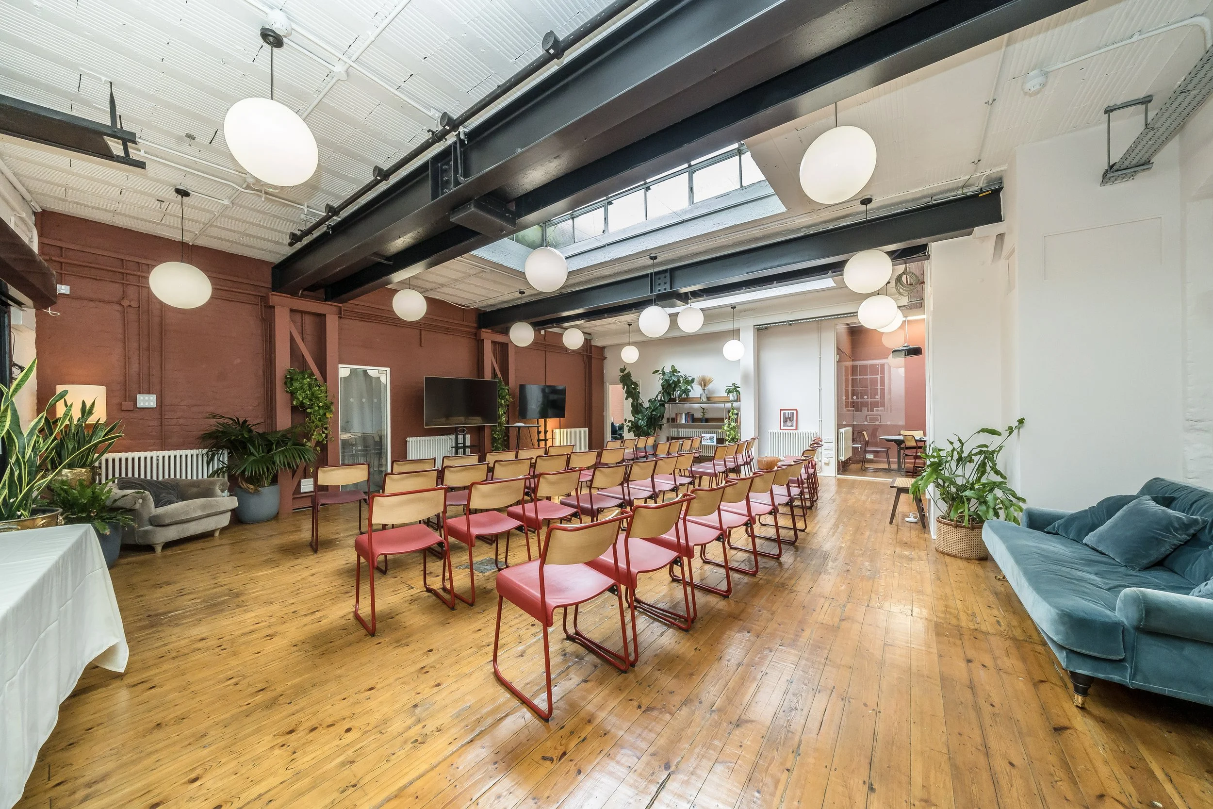 A spacious room with wooden flooring, decorated with plants and furniture, set up for a presentation or meeting with chairs arranged in rows facing two large screens at the front.