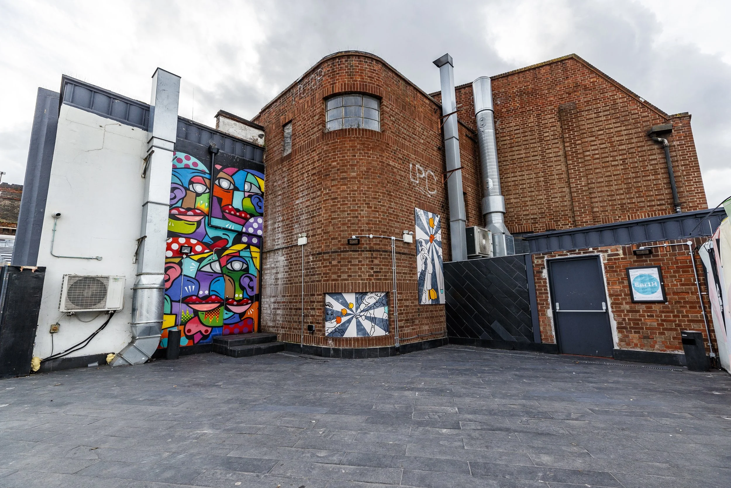 Brightly colored mural of abstract faces on the side of a brick building with gray sky above.