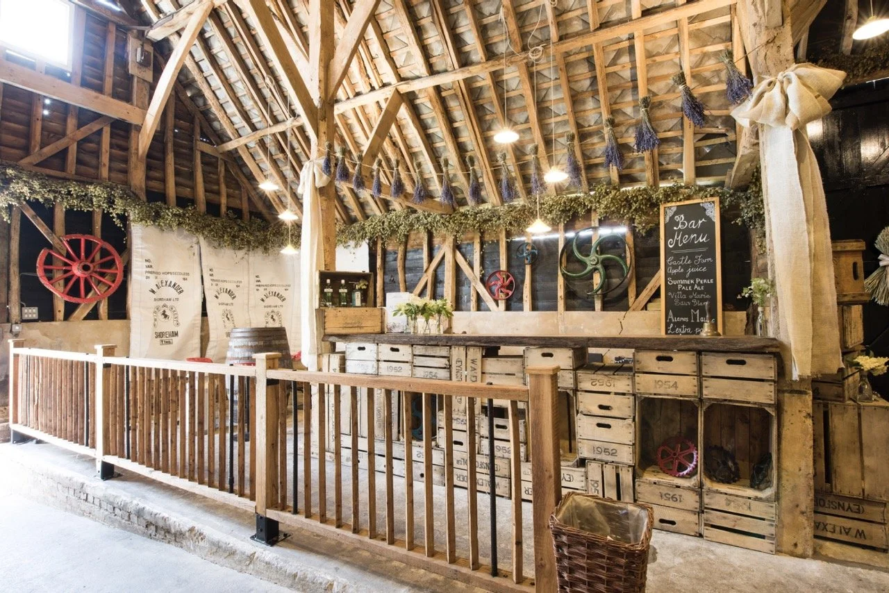 Rustic bar area with wooden crates and decorations, including lavender bundles and vintage wheels, inside a barn-like setting with wooden beams and a chalkboard menu.