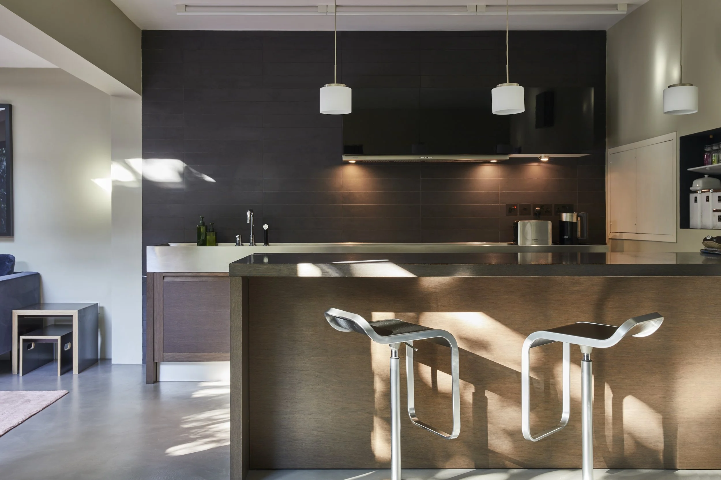 Modern kitchen with dark tile backsplash, wooden cabinetry, white pendant lights, stainless steel appliances, and a breakfast bar with two contemporary barstools.
