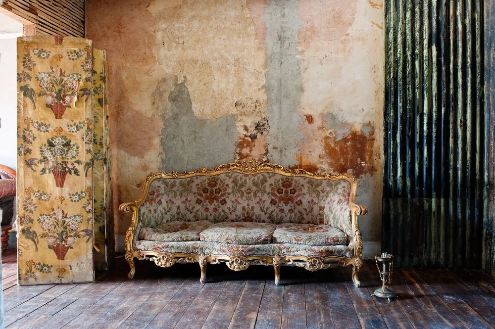 An ornate vintage sofa with floral upholstery against a weathered wall, next to a decorative folding screen with floral designs and a small metal candle holder on the floor.