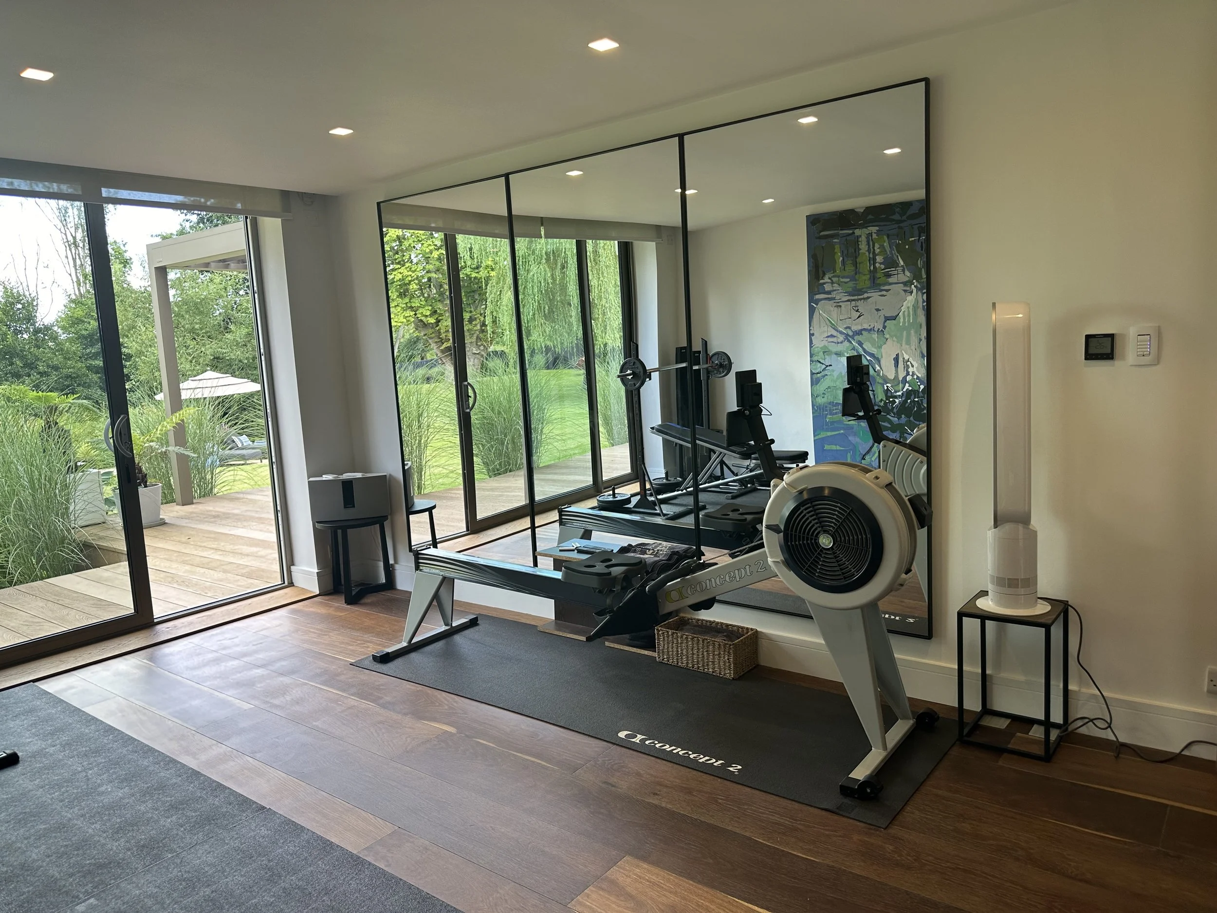 Home gym with a rowing machine, large mirror, sliding glass doors showing outdoor patio and greenery, small side table with a humidifier, and decorative wall art.