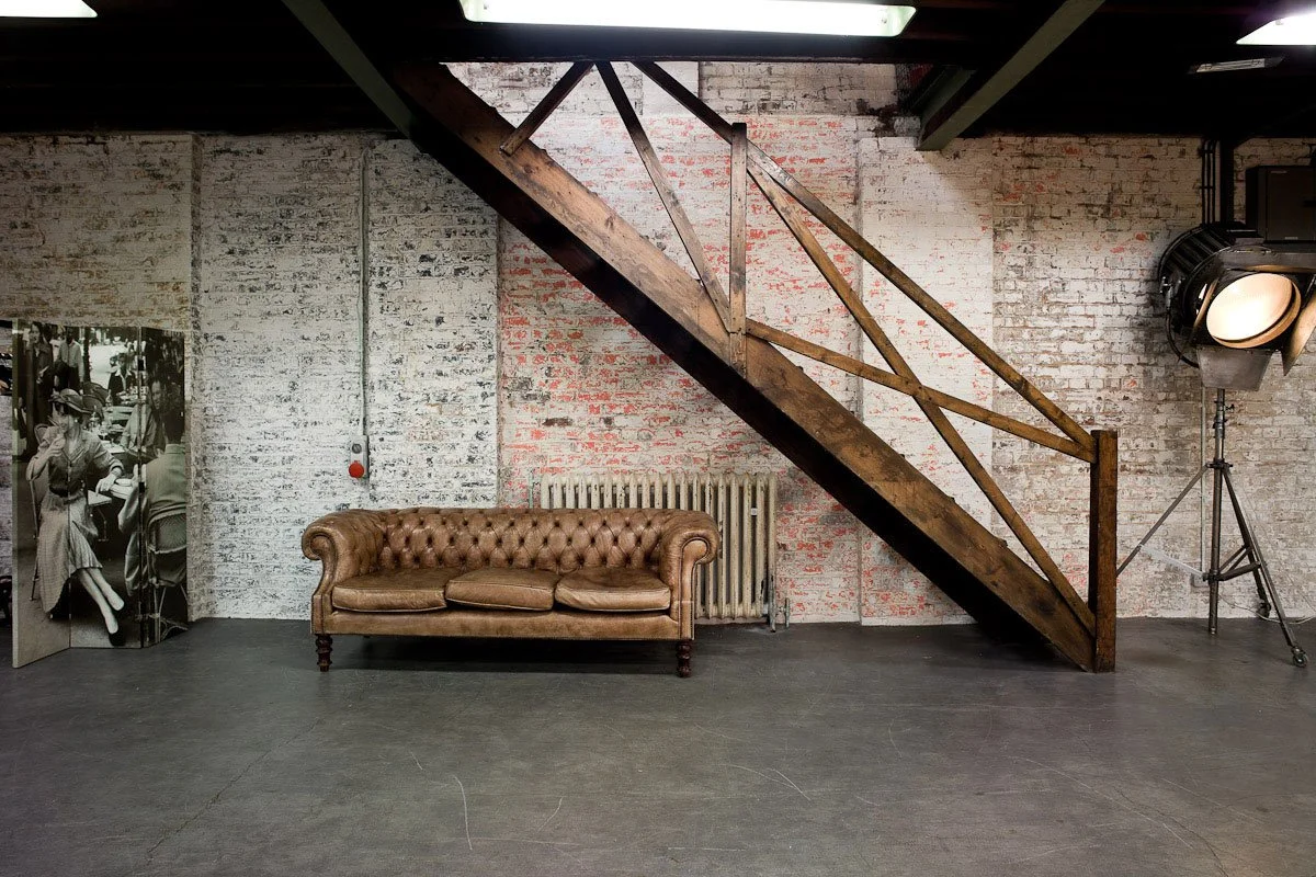 An industrial-style room with a brown leather tufted sofa, a staircase made of wood and metal, a black and white photo print leaning against the wall, and stage lighting equipment on a tripod.