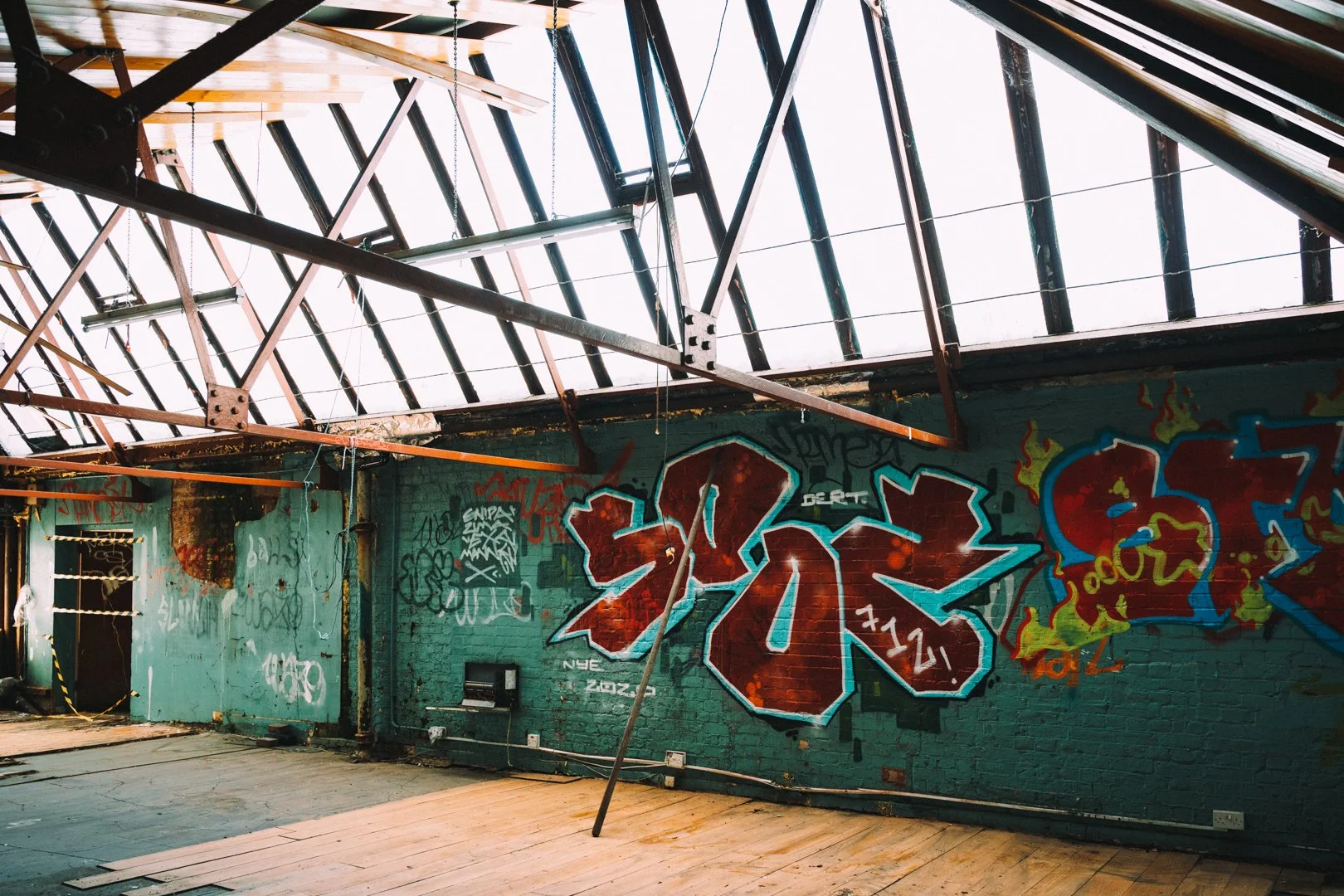 Interior of an abandoned building with graffiti on brick walls, a damaged roof with exposed beams, and broken wooden flooring.
