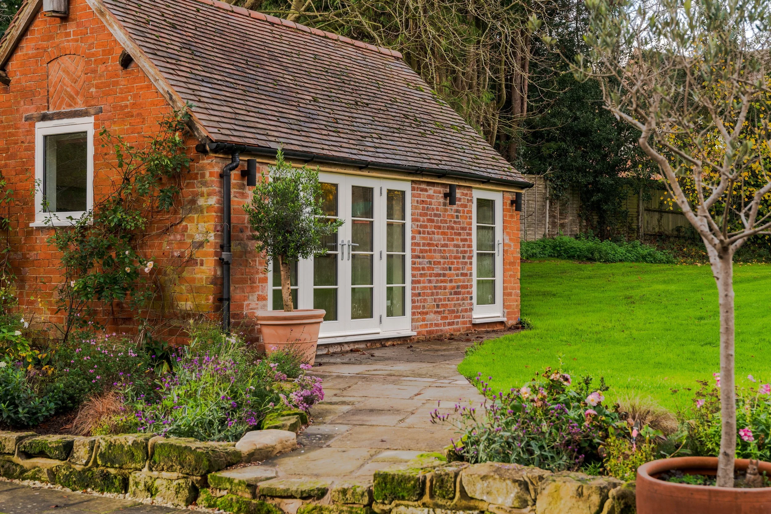 A brick house with a sloped roof, glass double doors, and windows. The house has a garden with potted plants, flowers, and a paved stone pathway leading to the entrance. There is a well-maintained lawn and trees in the background.