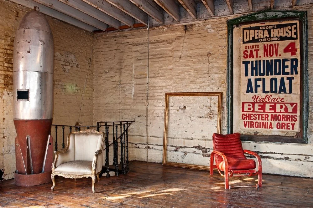 Vintage interior with a large rocket, an ornate armchair, an empty picture frame, a red lounge chair, and a theater poster on a worn brick wall.