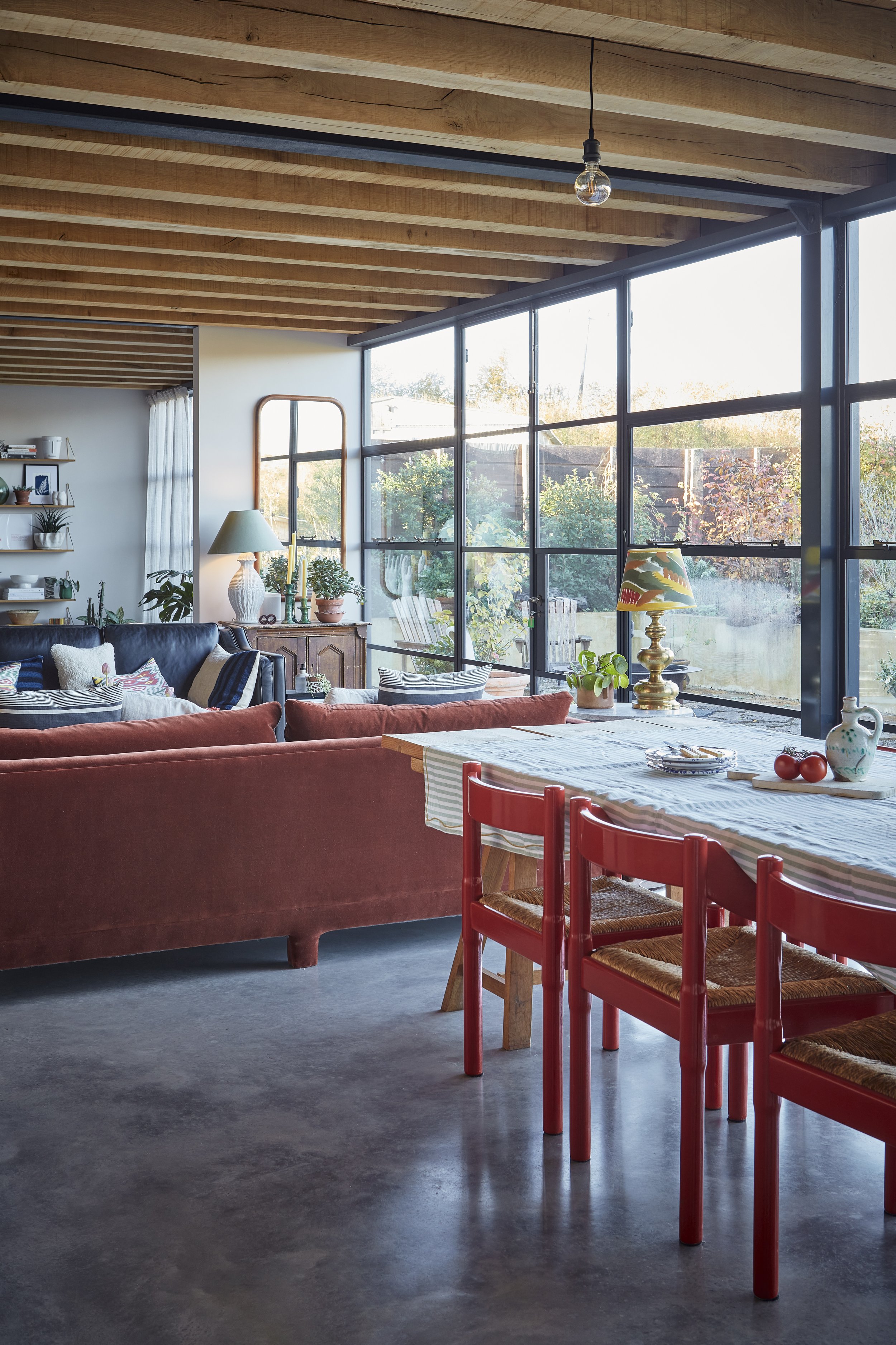 A spacious living and dining area featuring a high wooden ceiling, large windows letting in natural light, a dark leather sofa with patterned pillows, a wooden sideboard with a lamp and plants, a wall-mounted shelf with decor, and a dining table with