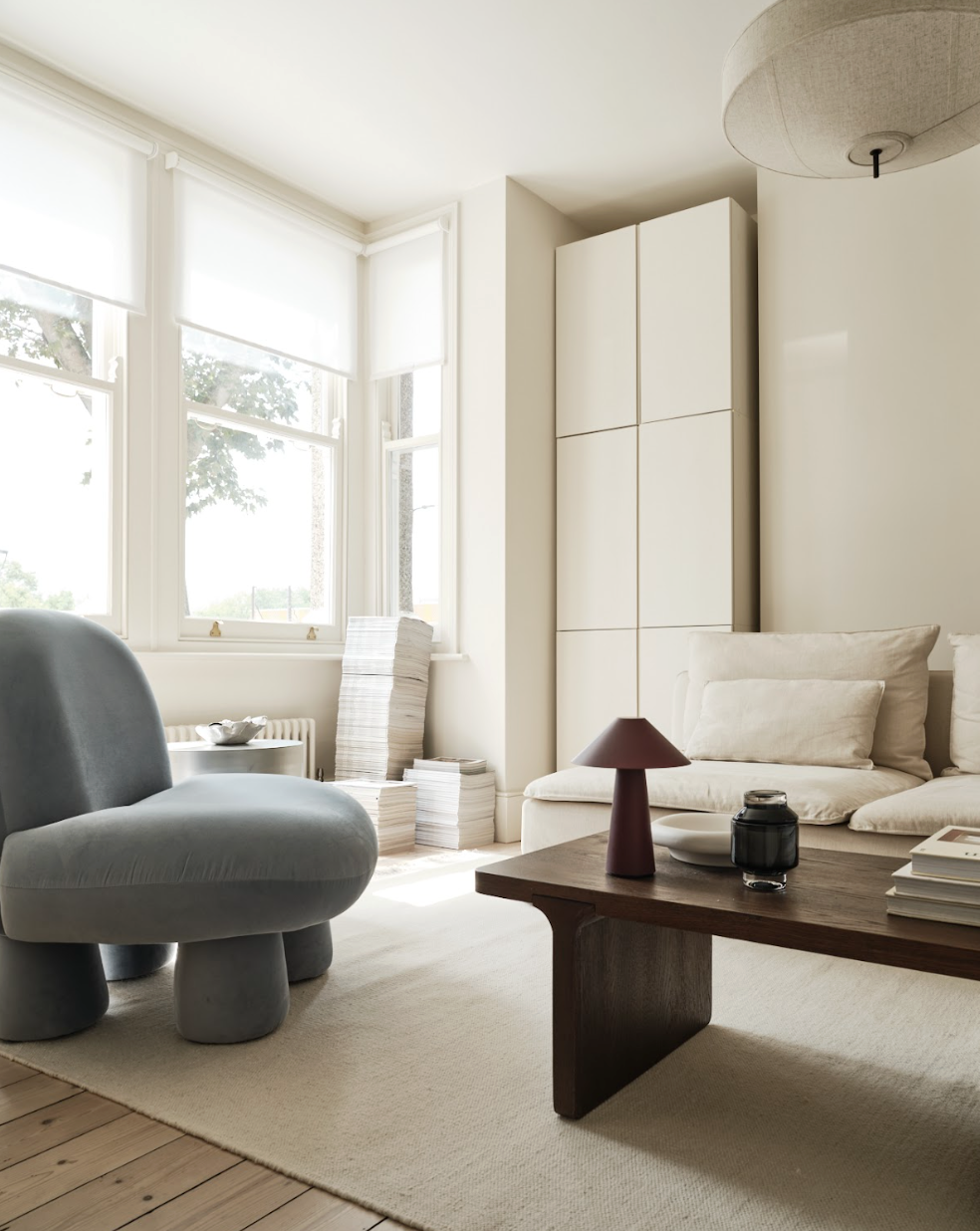 Living room with large windows, white walls, beige sofa with cushions, grey chair, wooden coffee table with a lamp, candle, and books, and a white cabinet in the corner.