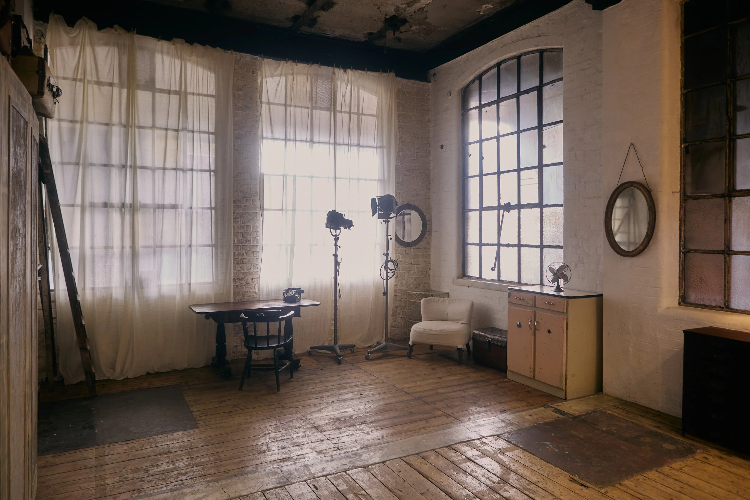 An interior room with large industrial-style windows, white brick walls, wooden floors, and simple furniture, including a small table with a black chair, two studio lights, a white armchair, and a vintage cabinet.