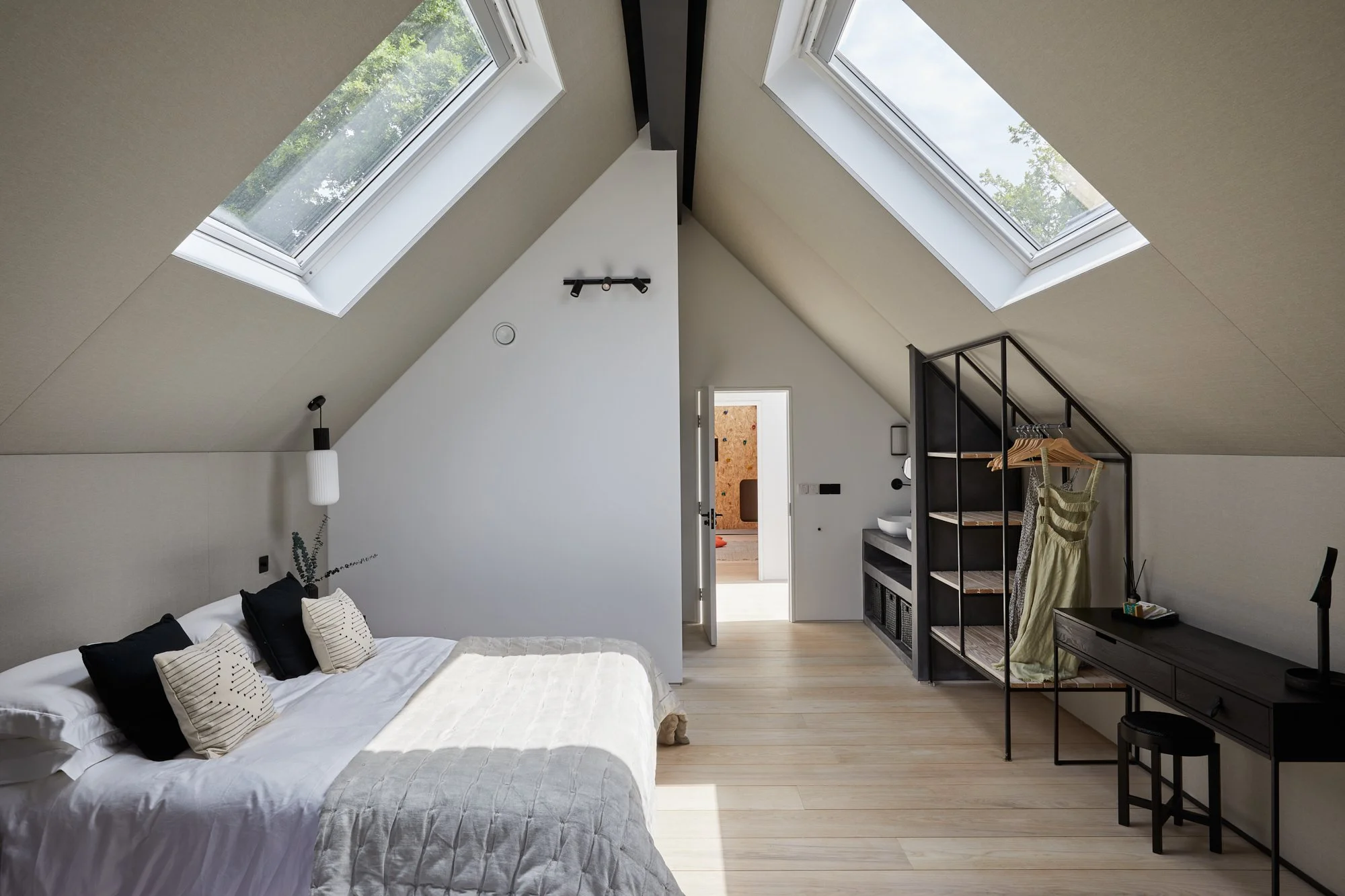 A minimalist bedroom with a sloped ceiling featuring two skylight windows. The room has a cozy bed with black and beige pillows, a wall-mounted lamp, a small black desk with a chair, and a black metal shelving unit with clothes hanging on it. Light w