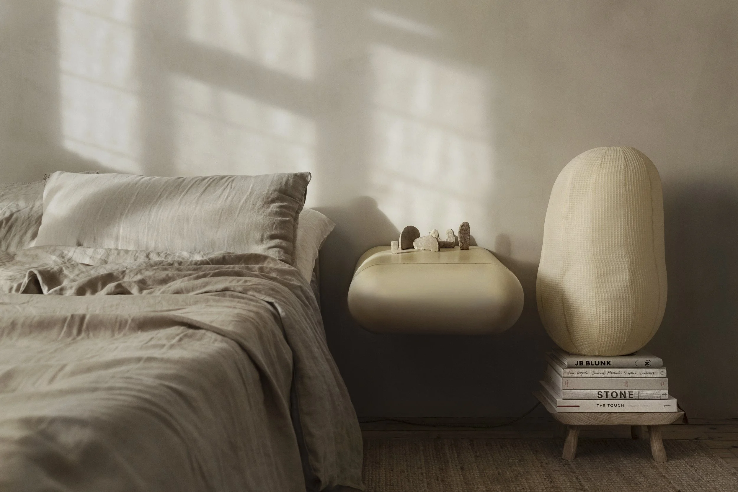 A cozy bedroom corner with a bed with beige linens, a floating nightstand with stones on top, a stack of books with a large textured beige vase or sculpture on top, and sunlight casting shadows through a window.