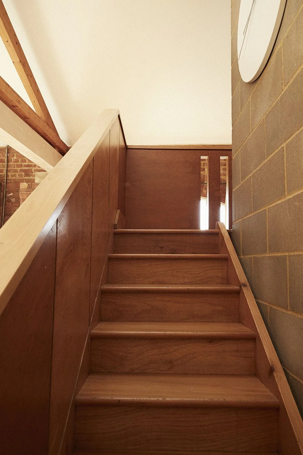 Wooden staircase leading to an upper level, with a brick wall on the right and a ceiling with exposed wooden beams.