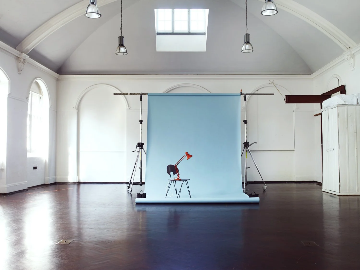 Empty photography studio with a blue backdrop, a chair with a red desk lamp, and professional lighting equipment in a spacious room with wooden floors and white walls.